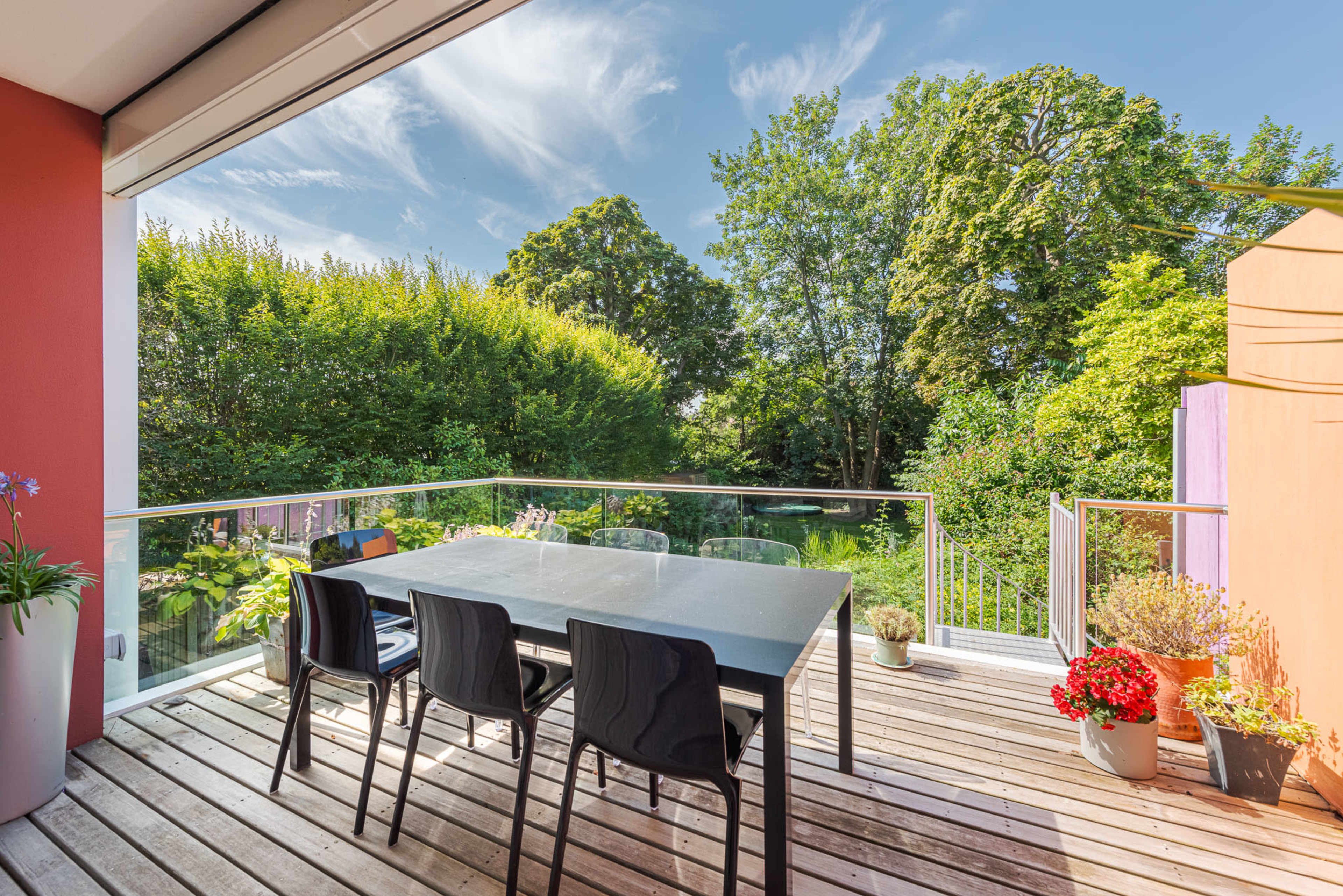A modern deck features a large table with black chairs, surrounded by greenery and colorful plants.