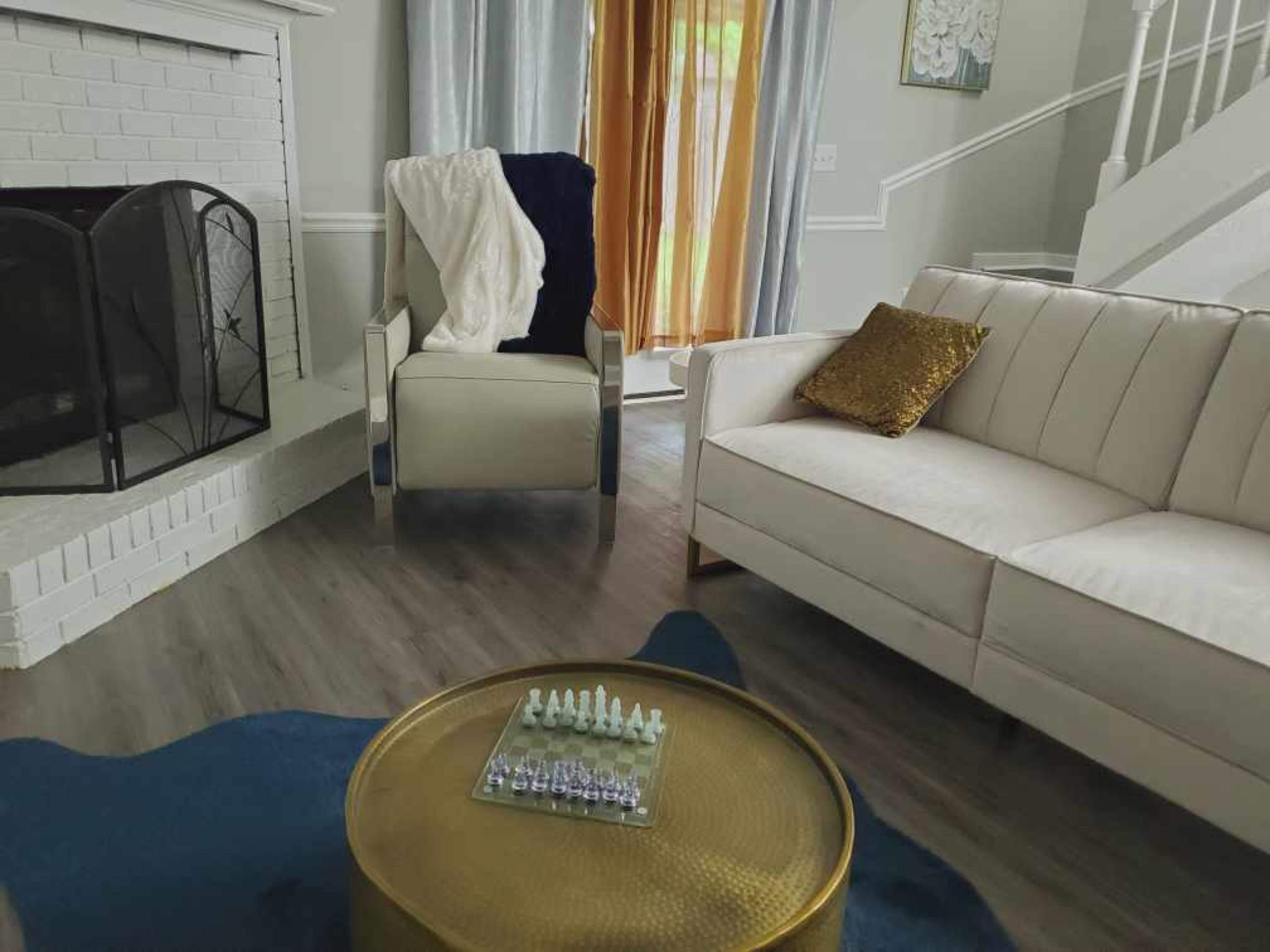 The living room features a white sofa, a decorative chair, a round gold coffee table with a chess set, and large windows with sheer curtains.