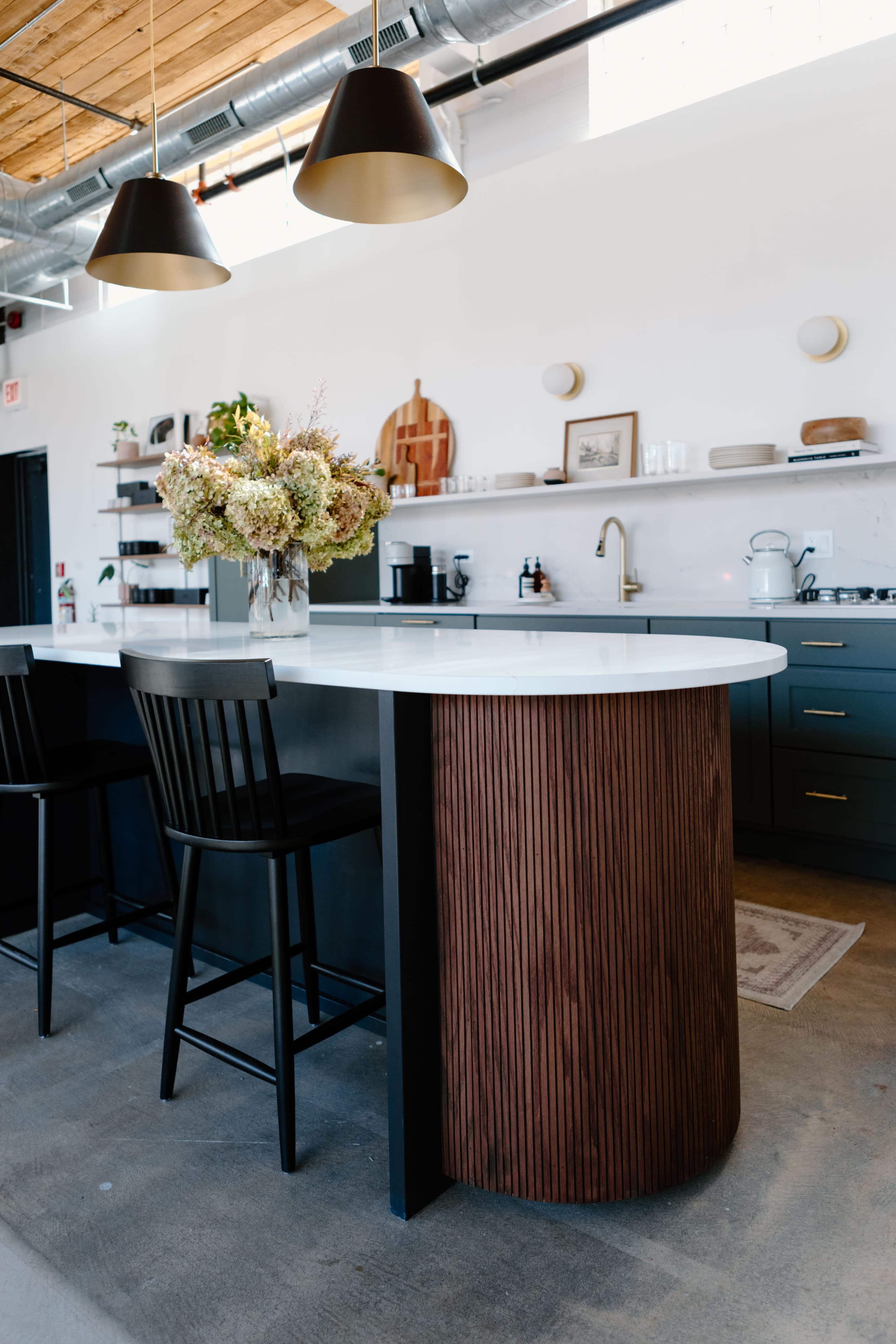 A modern kitchen featuring a curved wooden island with black bar stools, a white countertop, and neutral decor.
