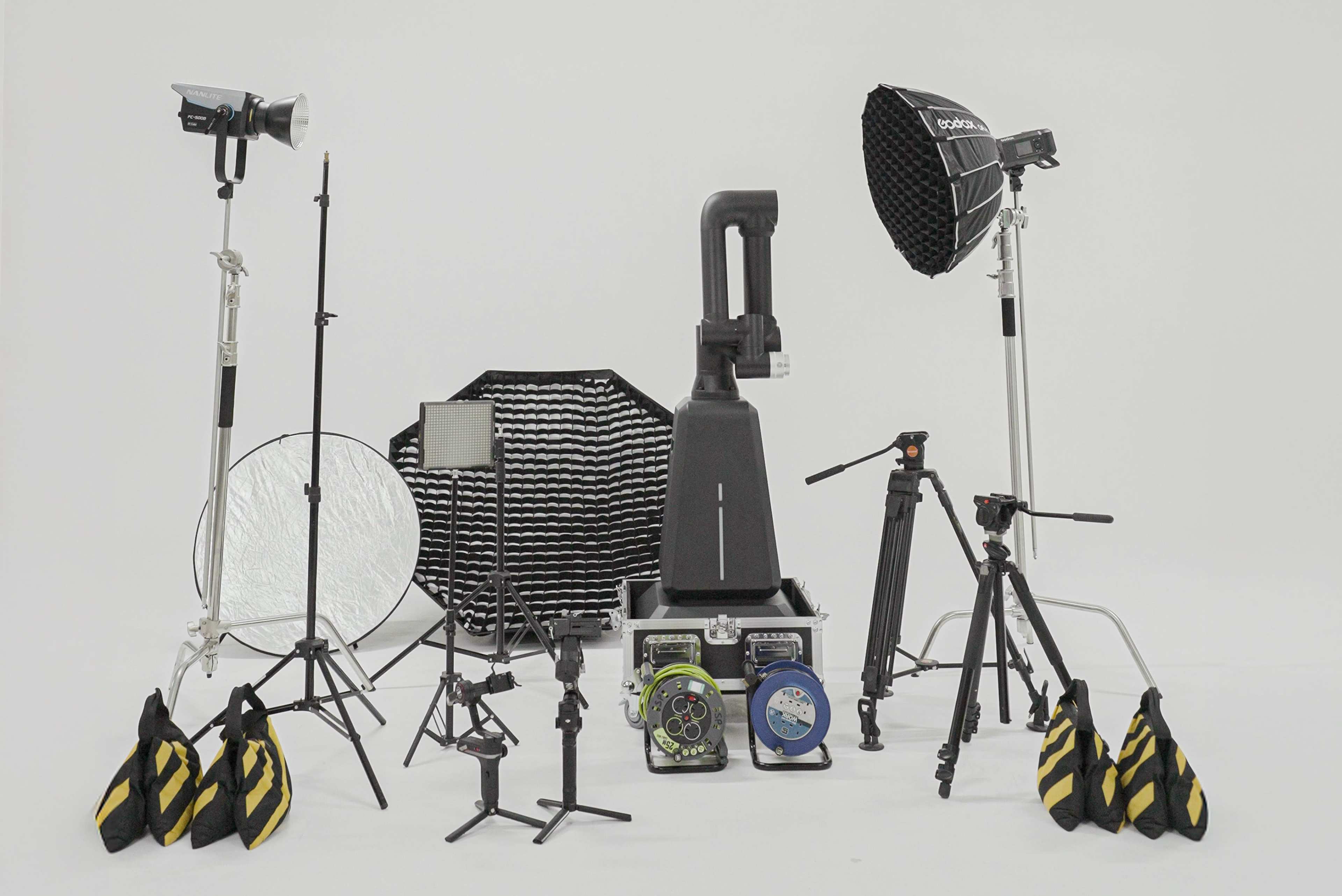 The image shows a collection of photography and lighting equipment arranged on a white background, including tripods, softboxes, reflectors, and various lights.