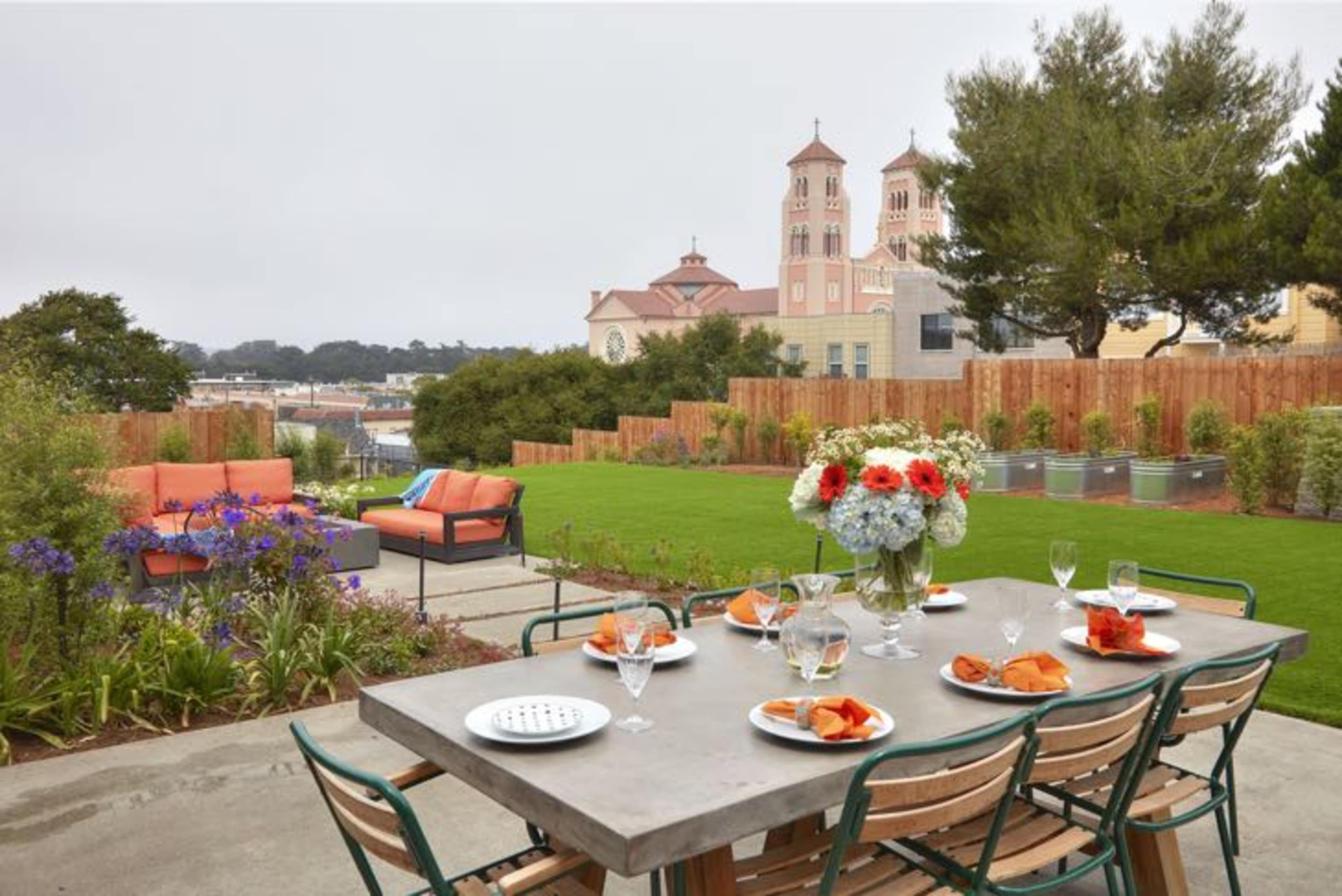 A dining table is set in a landscaped backyard with flowers and a view of a building with two towers in the background.