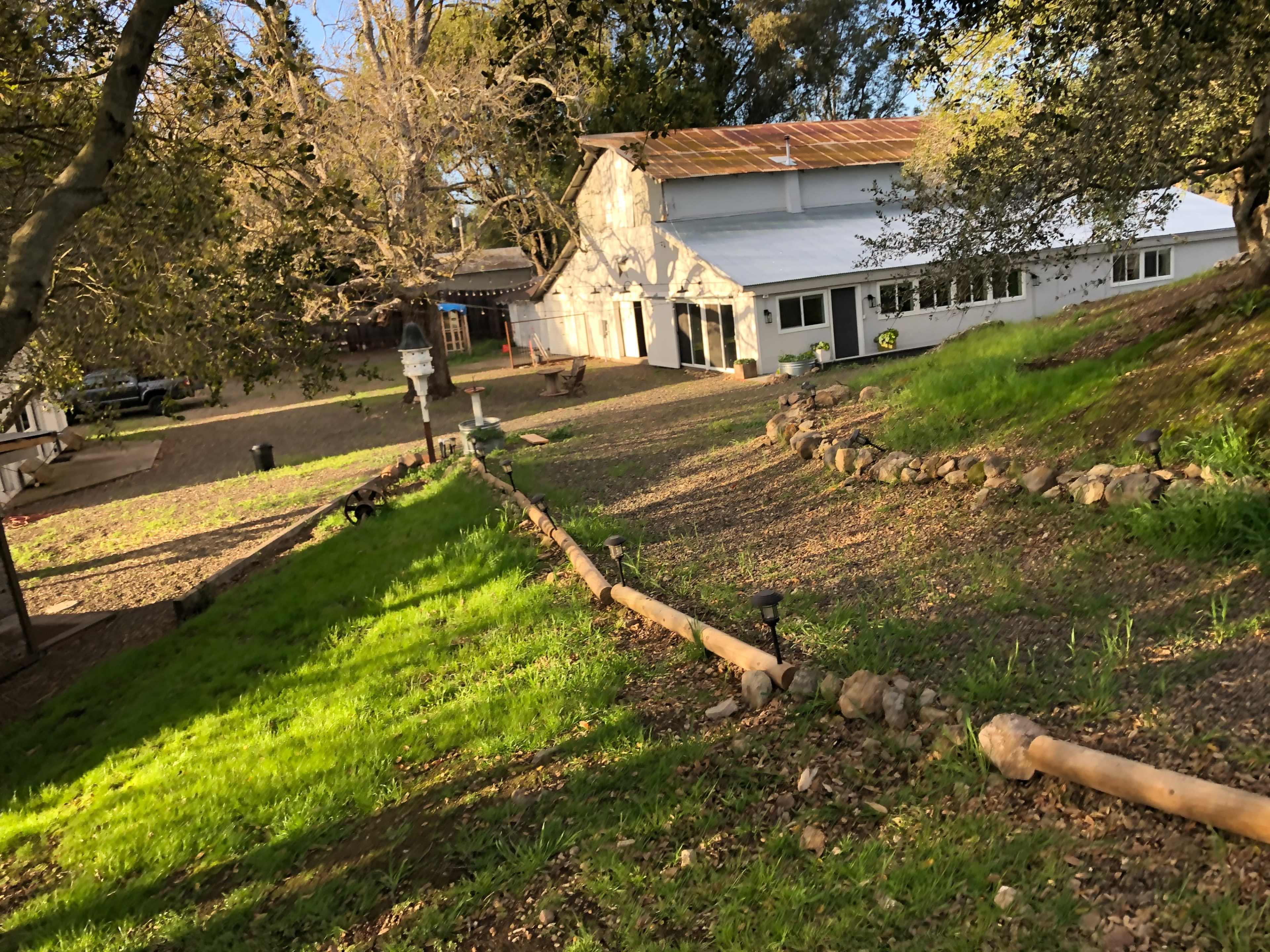 A rustic building with a metal roof is situated in a green, sloped yard surrounded by trees and a gravel path.