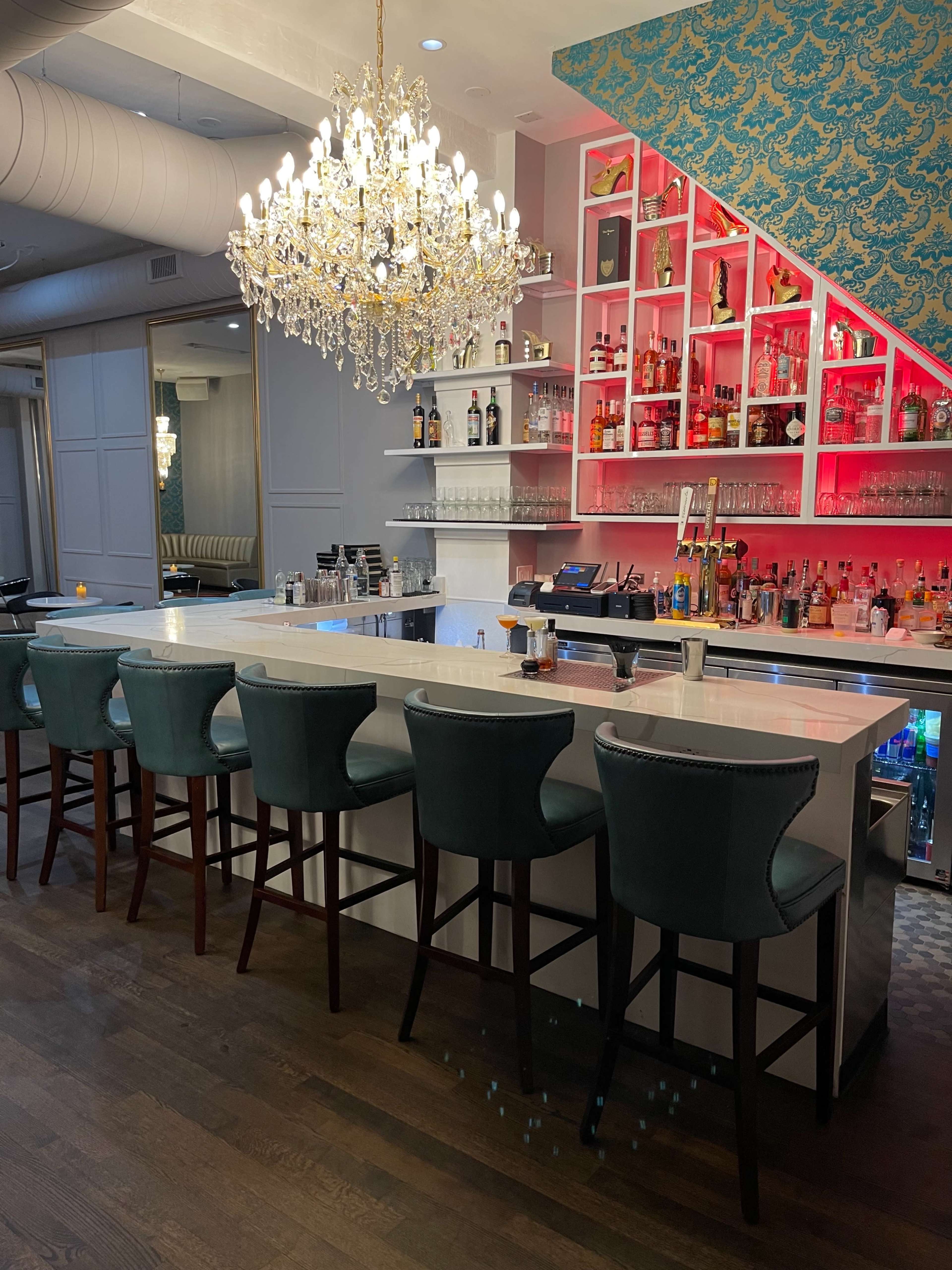 An elegant bar area featuring a chandelier, a backlit shelf with various bottles, and a row of teal chairs at a white countertop.