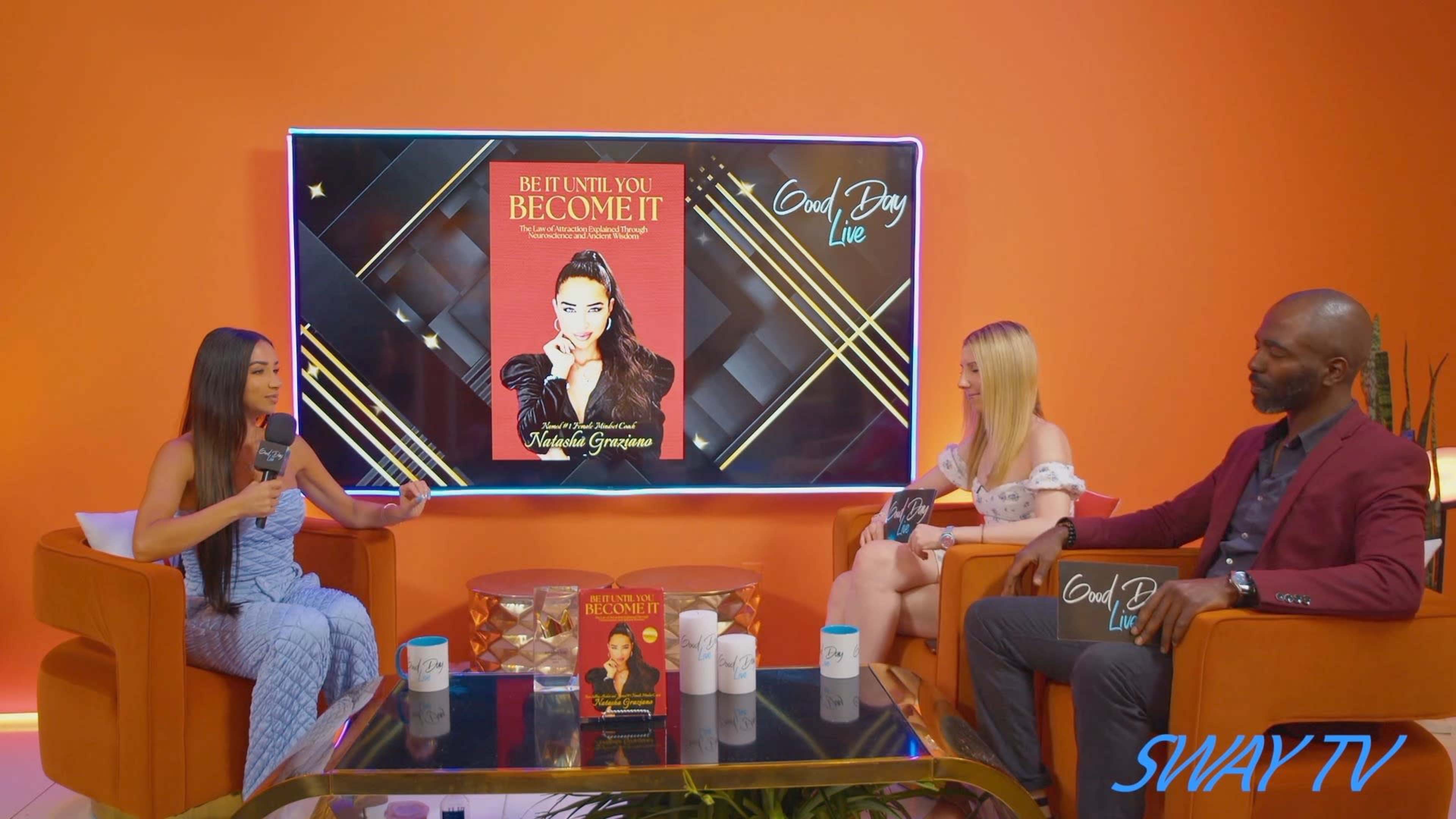 A talk show set features three hosts seated with a backdrop displaying a book titled "Be It Until You Become It."