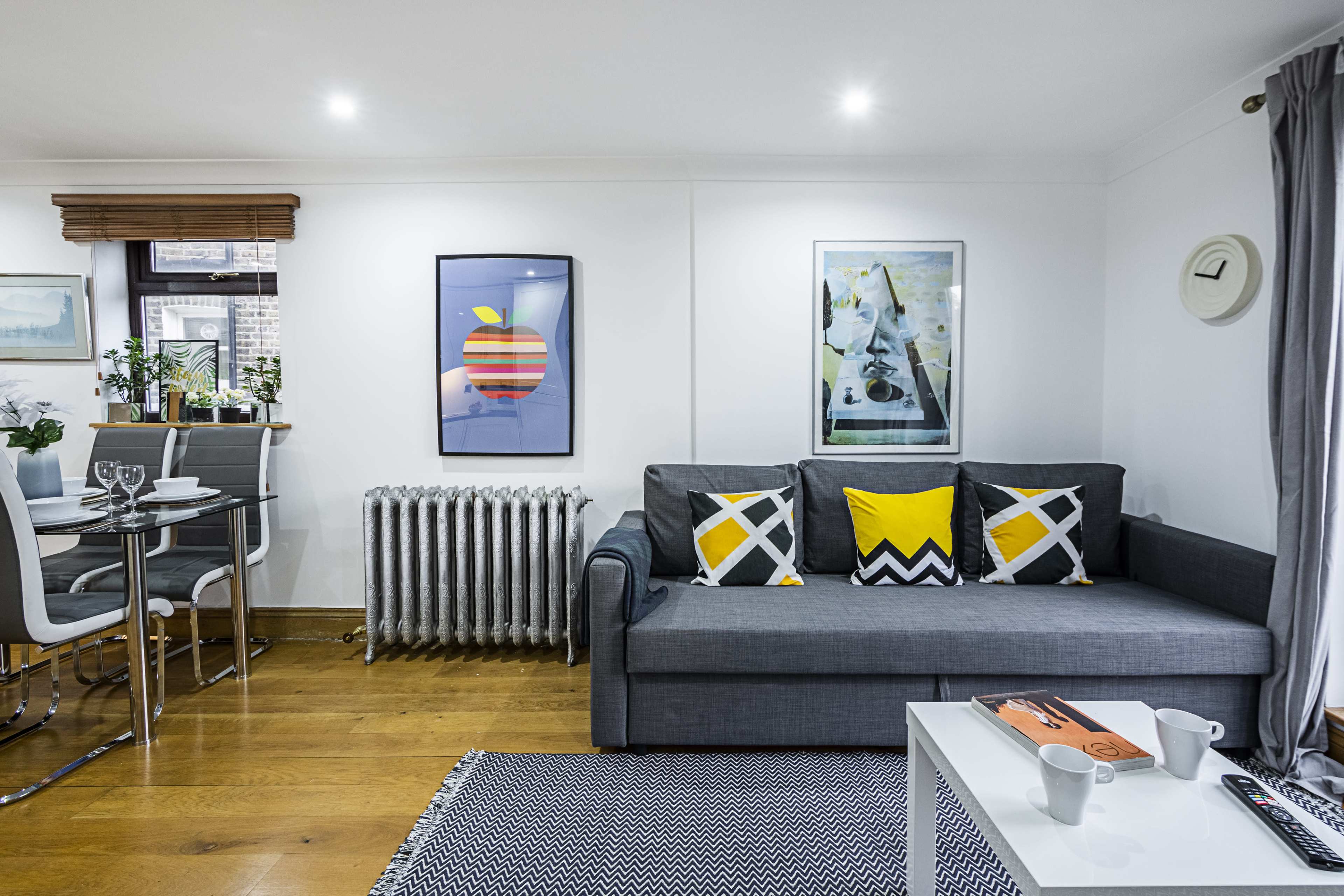 A cozy living space with a gray sofa adorned with yellow and black patterned cushions, a dining area with a small table set for two, and a textured rug on a wooden floor.