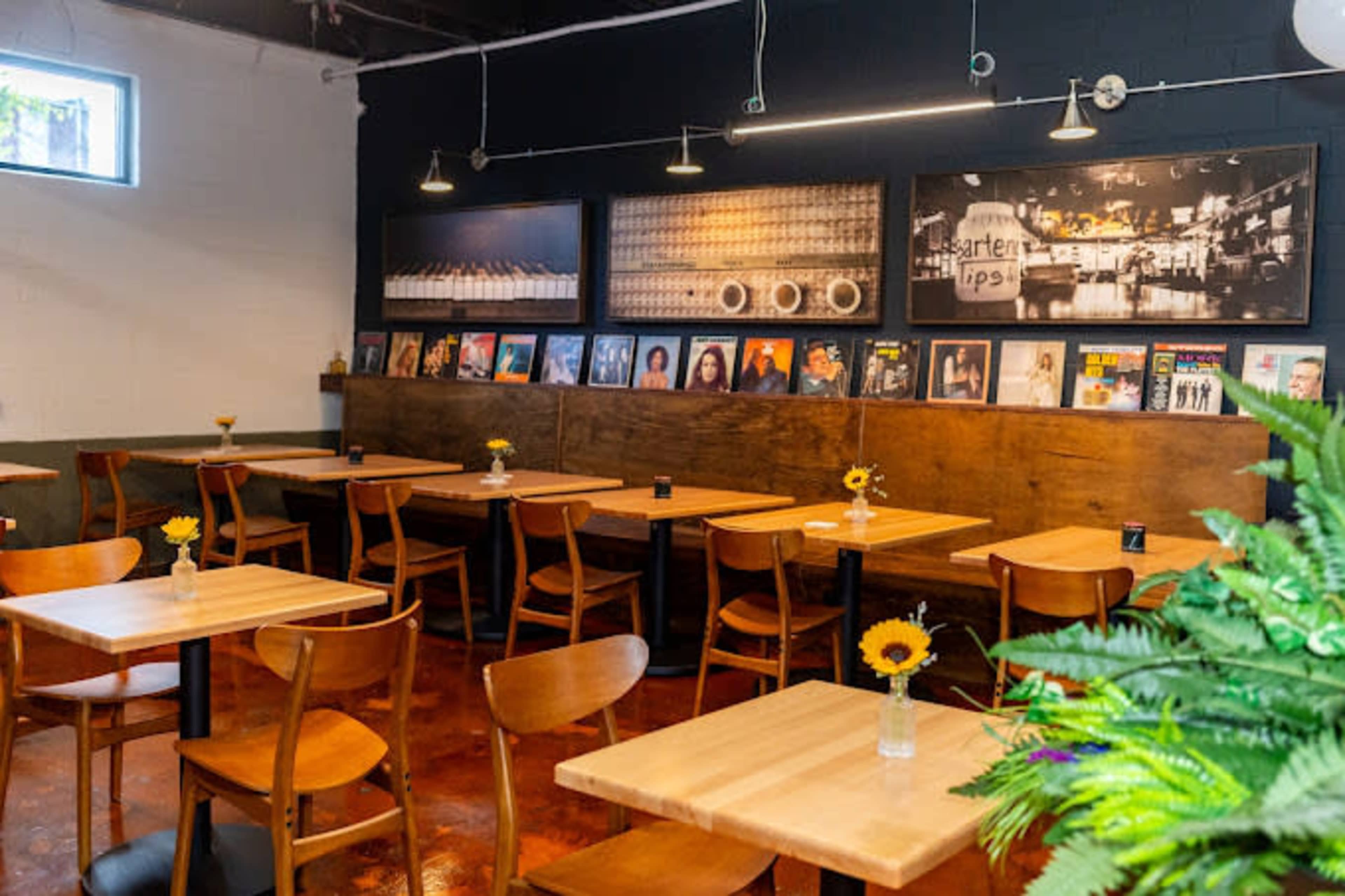 The image shows a restaurant interior with wooden tables and chairs, featuring artwork on the walls and small vases with flowers on each table.