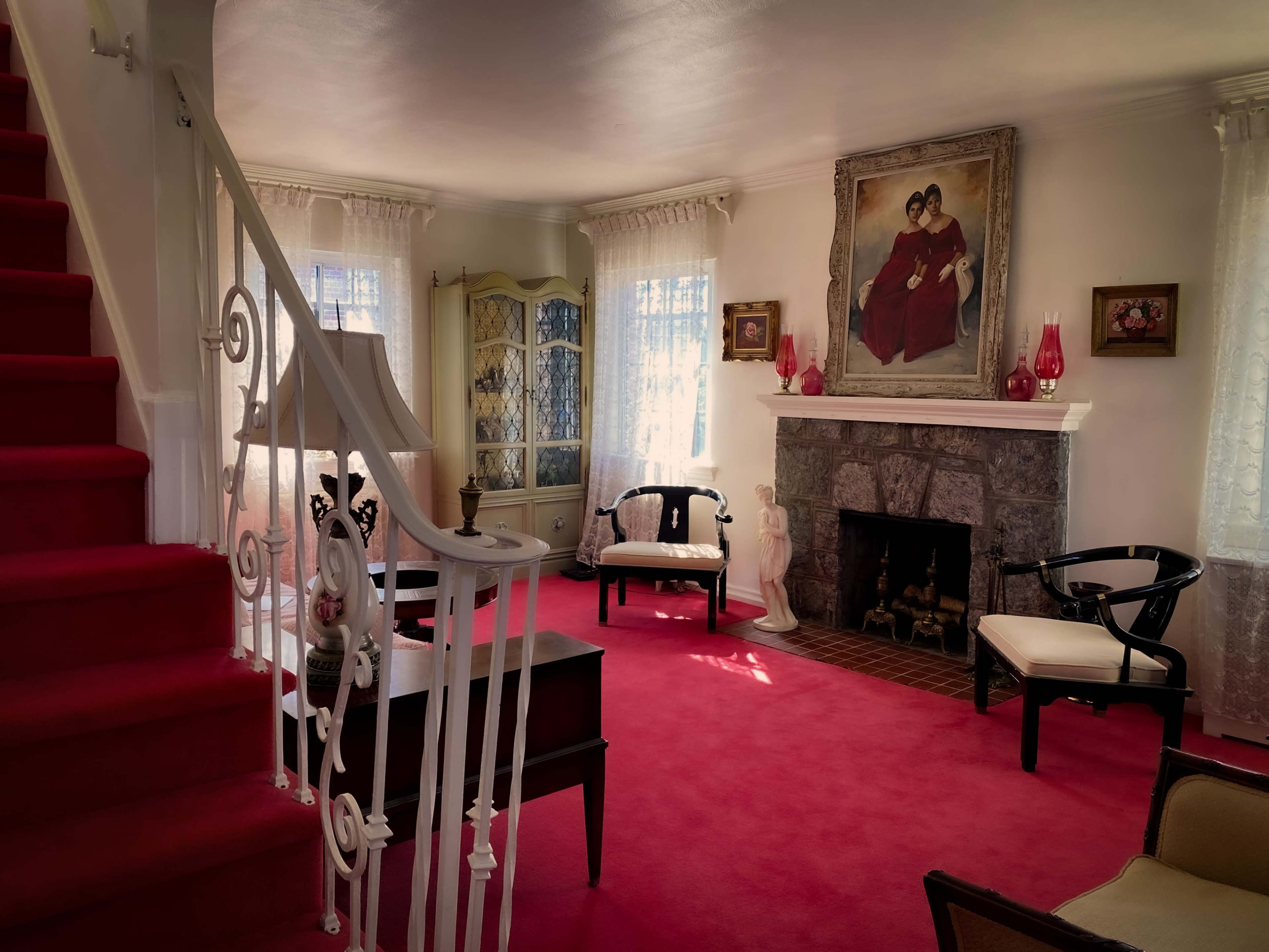 The image shows a brightly lit living room with red carpet, a staircase leading to the upper floor, a stone fireplace, and vintage furniture arranged around a central area, including a large painting on the wall.