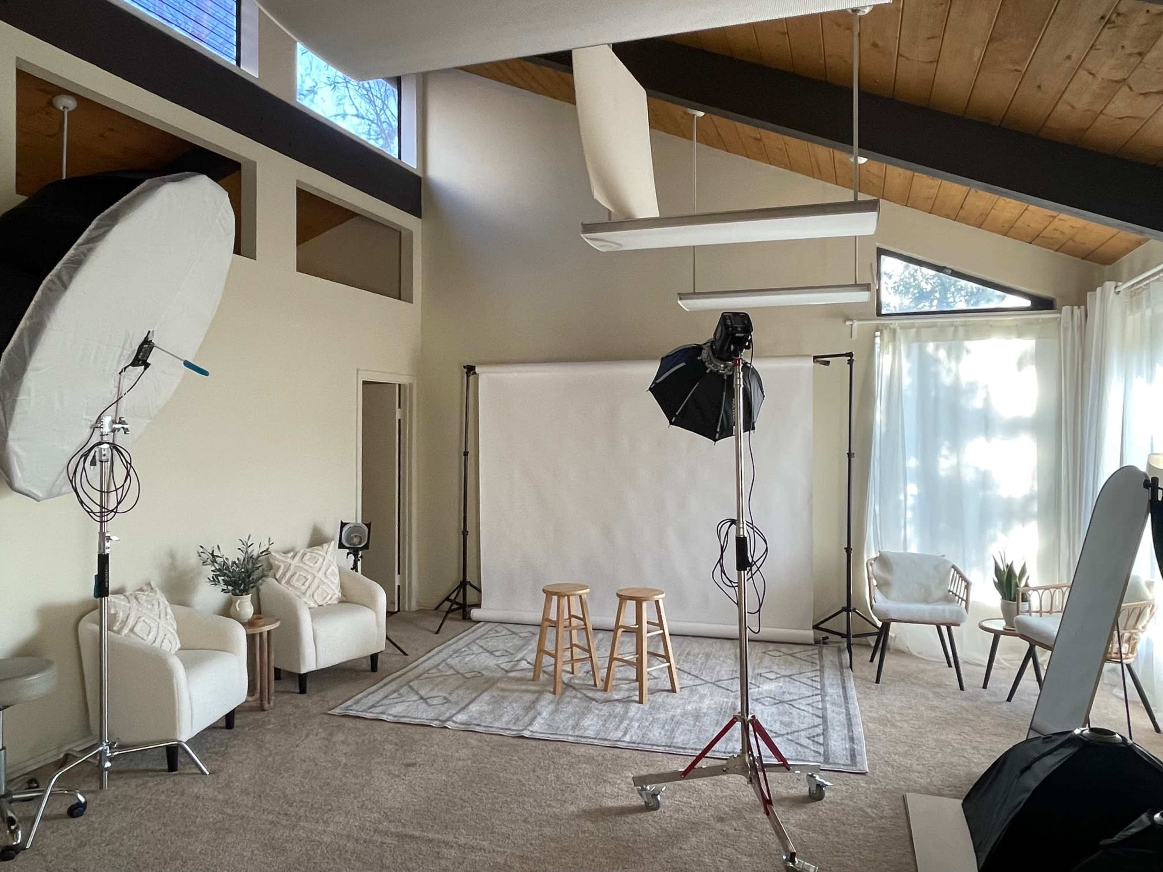 The image shows a photography studio featuring a light backdrop, two wooden stools, soft lighting equipment, and various seating arrangements.