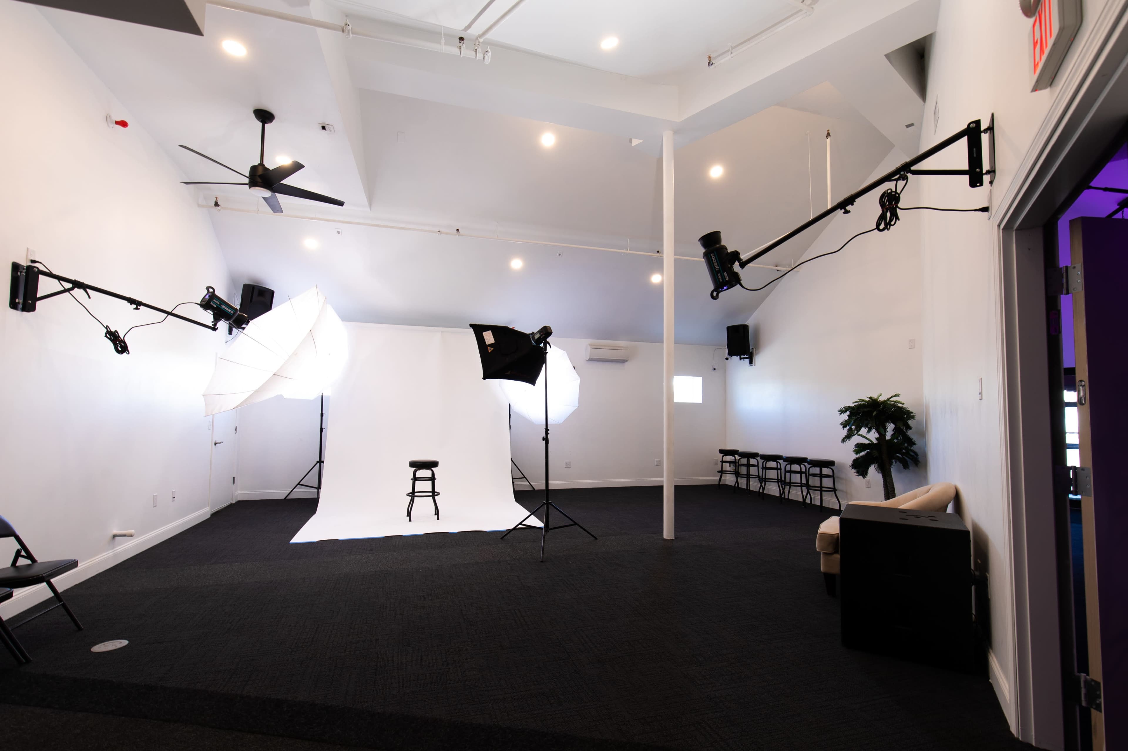 A photographic studio with backdrop equipment, lighting setups, and a single stool positioned in front of a white backdrop.
