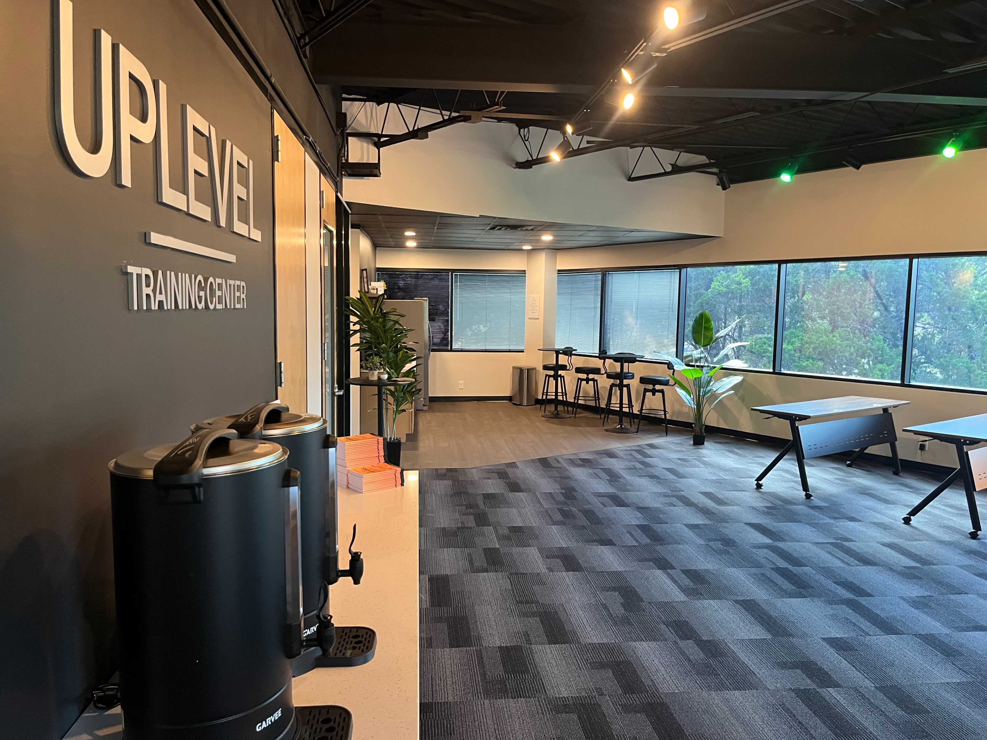 The image shows a modern training center with a clean layout, featuring a coffee station, tables, and large windows overlooking greenery.