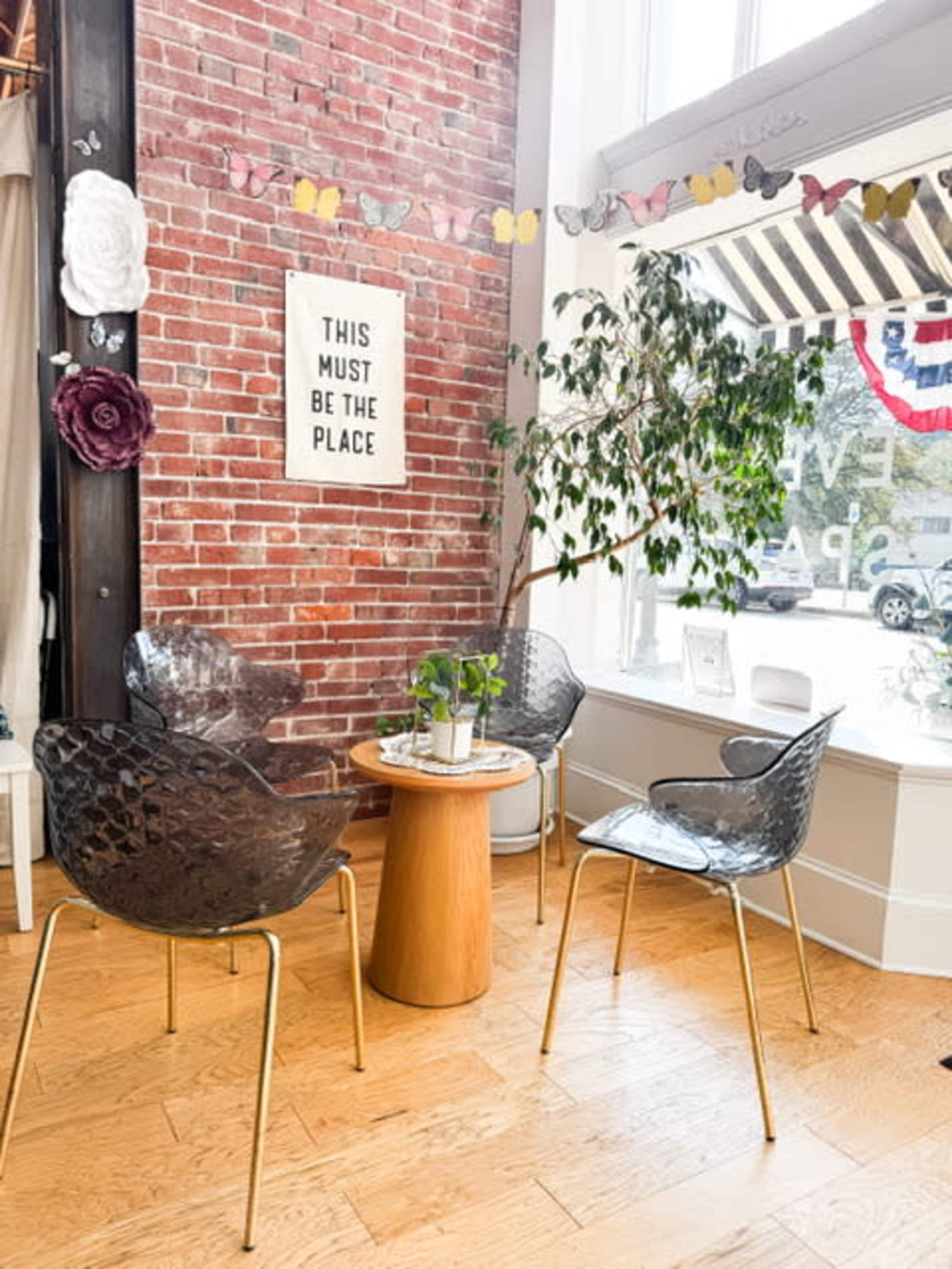 The image shows a cozy seating area with three transparent chairs around a wooden table, positioned against a brick wall with decorative elements and a window displaying a storefront.