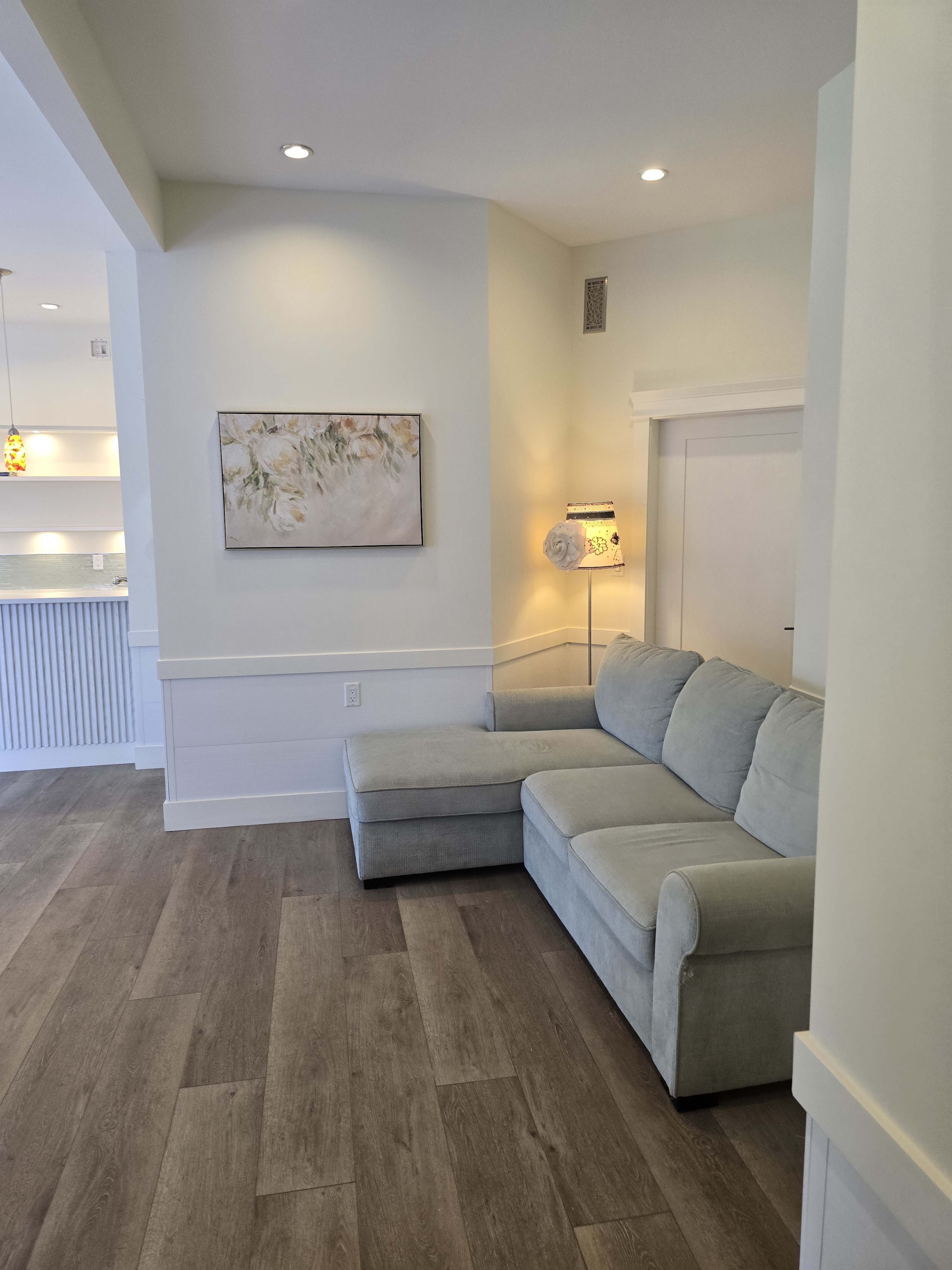 The image shows a light-colored living room with a sectional sofa, a framed floral painting on the wall, and a floor lamp beside the sofa.