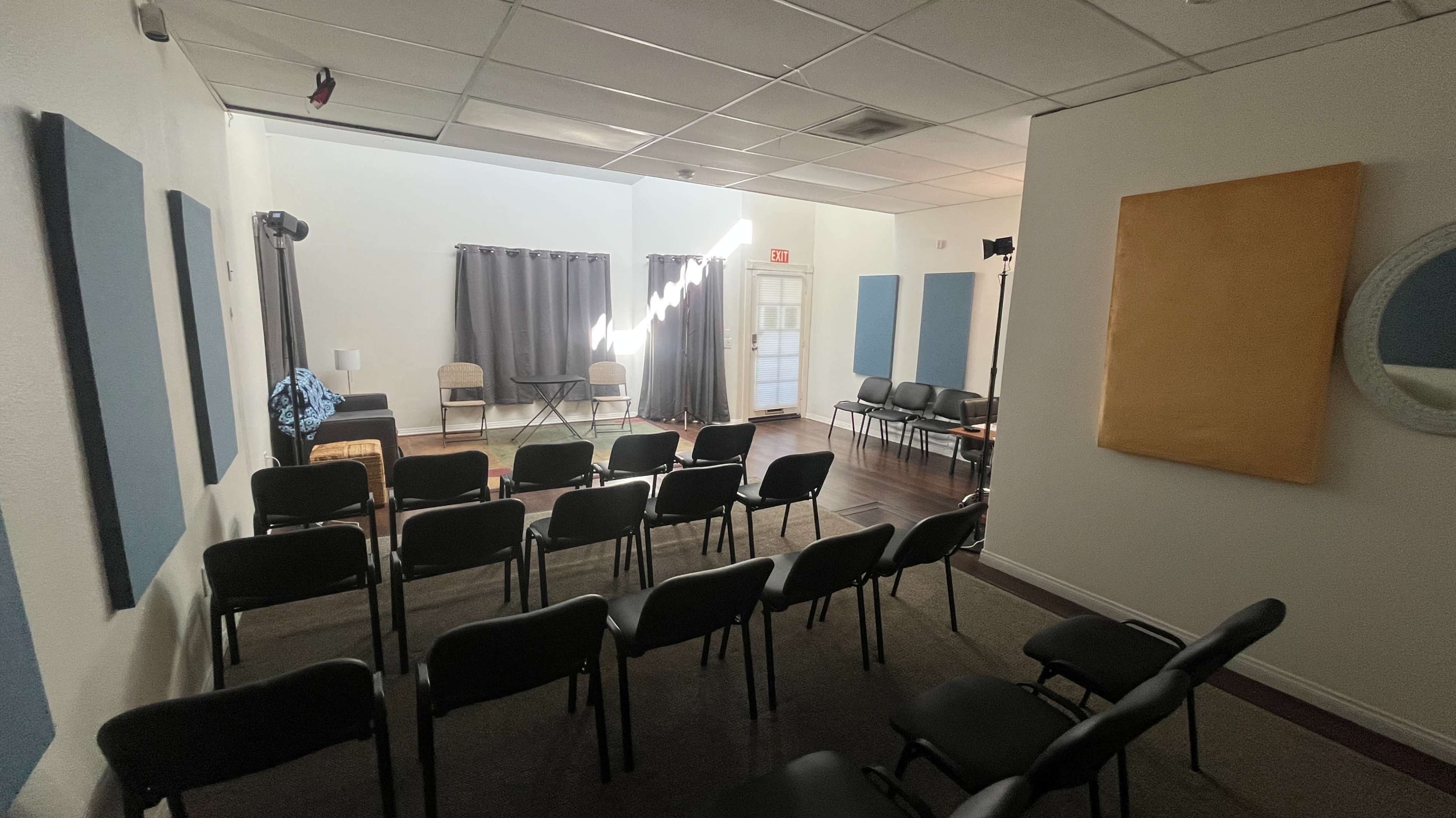 The image shows a sparsely furnished room with several rows of black chairs facing an empty performance area, featuring a small table and two chairs, with light streaming in through partially drawn curtains.