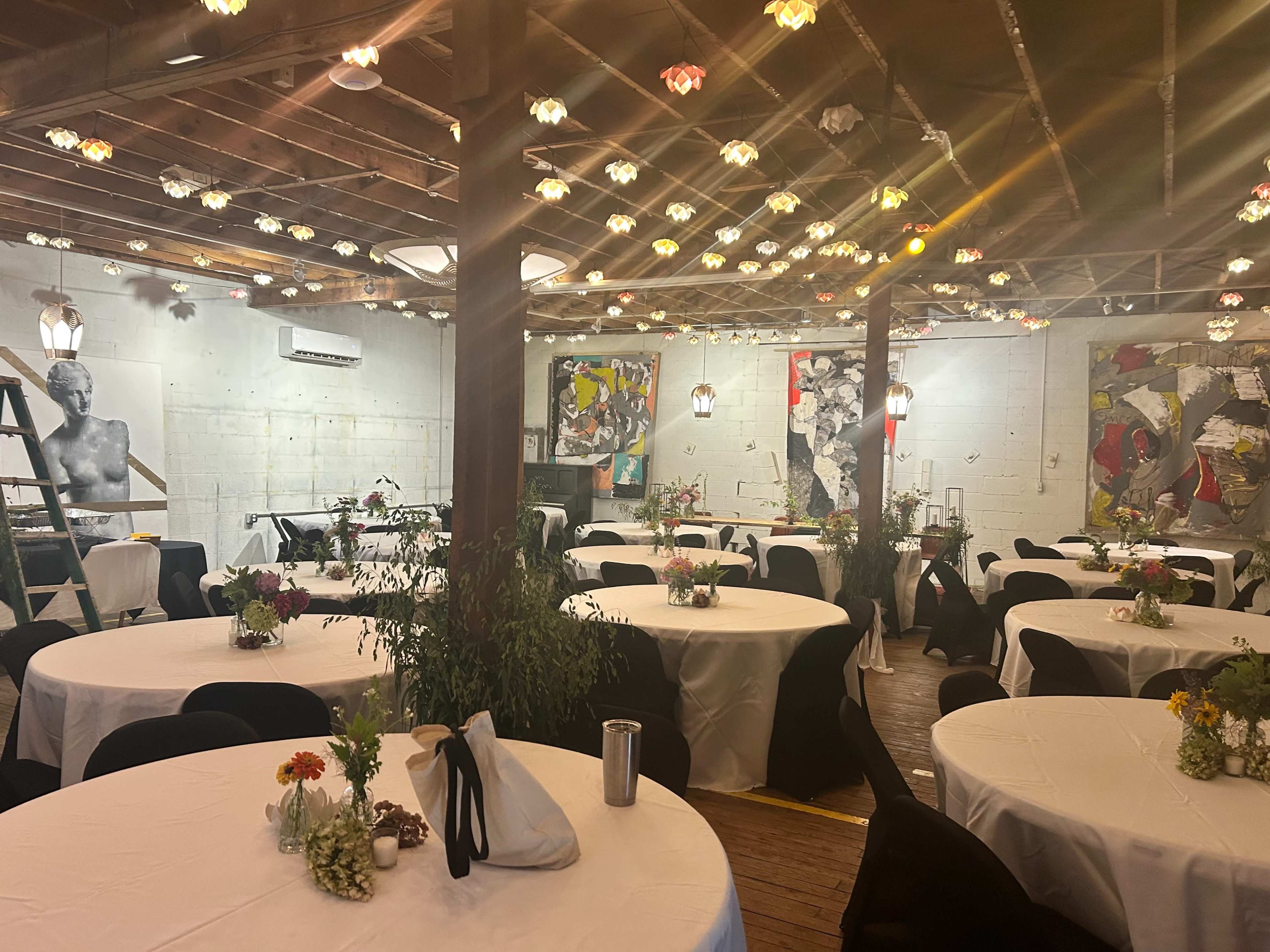 A decorated event space features round tables with white tablecloths arranged under a ceiling adorned with pendant lights and artwork on the walls.