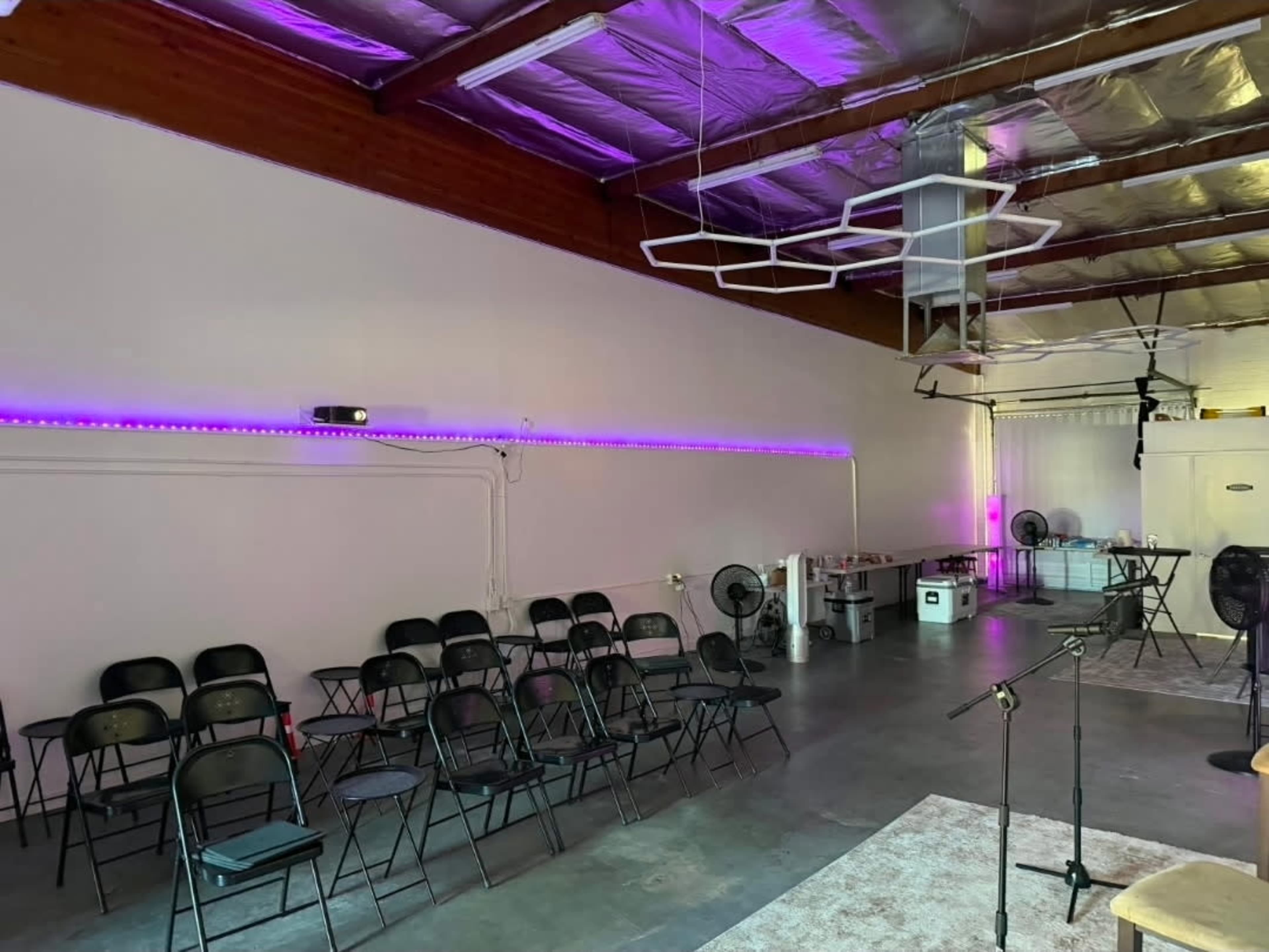 The image shows a large indoor space with rows of black folding chairs set up for an event, along with purple LED lighting along the walls and a small stage area.