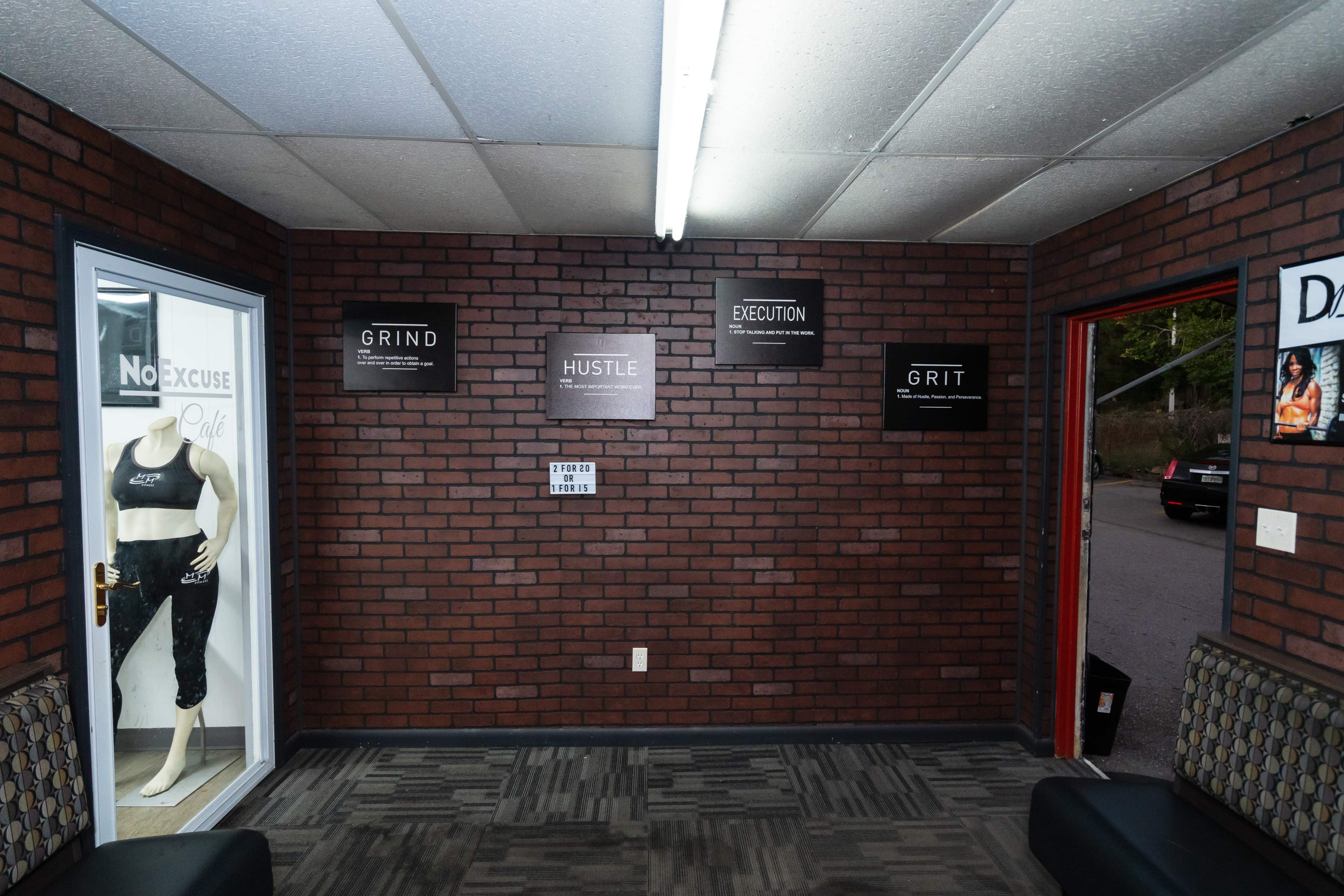 A brick-walled entrance area with a mannequin displaying athletic wear and motivational signs that read "GRIND," "HUSTLE," "EXECUTION," and "GRIT."