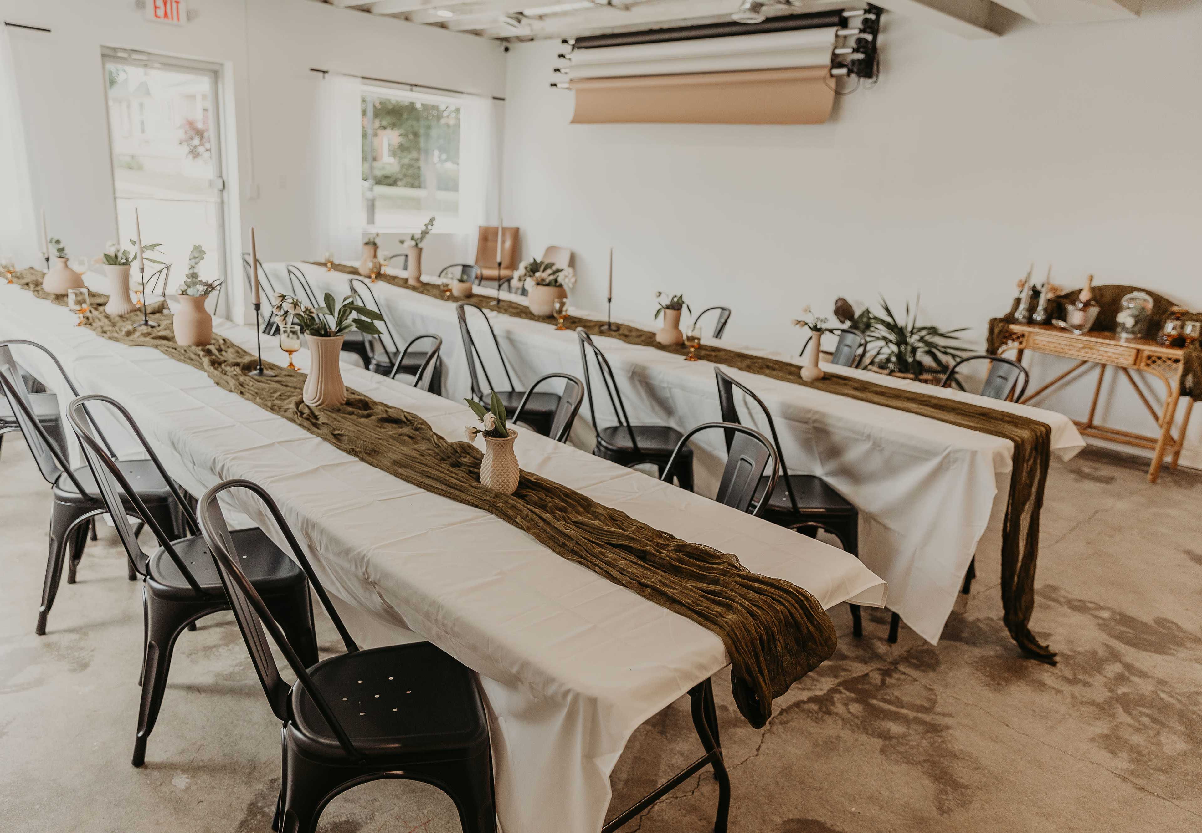 A large, empty room features two long tables covered with white cloths, adorned with green runners and centerpieces in vases, with black chairs arranged around them.