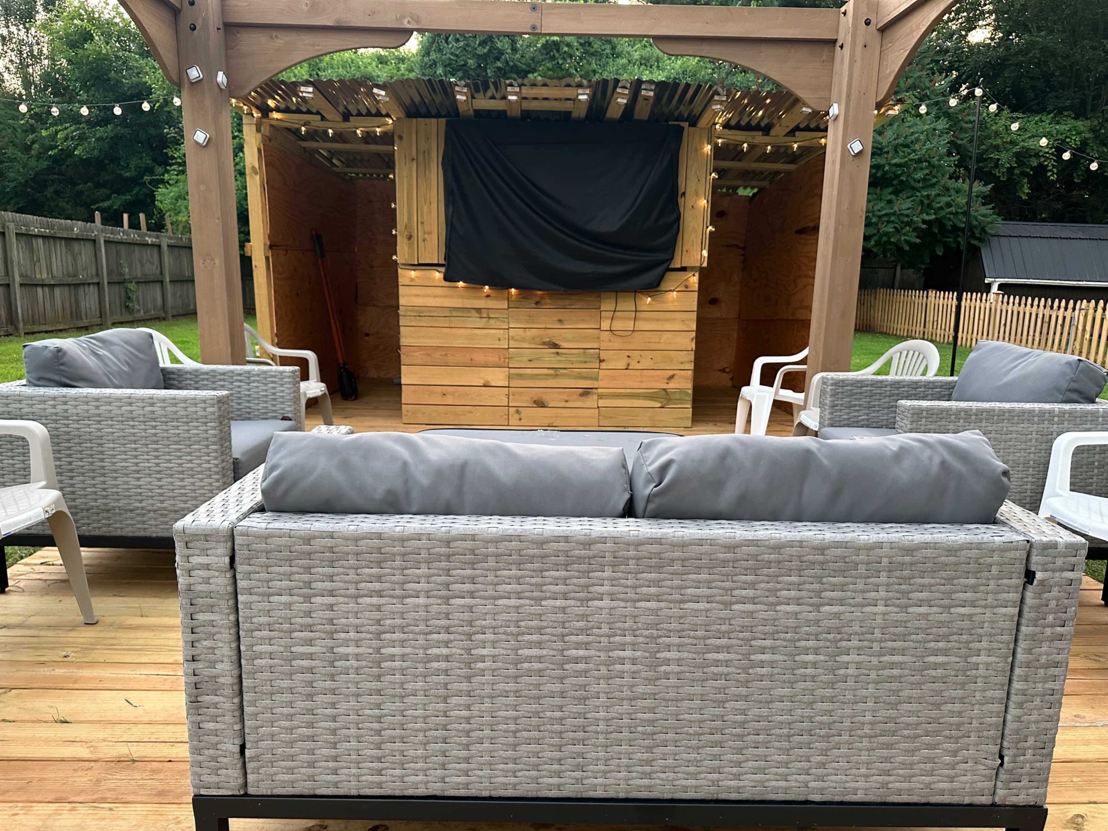 A patio area with several outdoor seating arrangements facing a wooden stage covered by a black cloth, illuminated by string lights overhead.