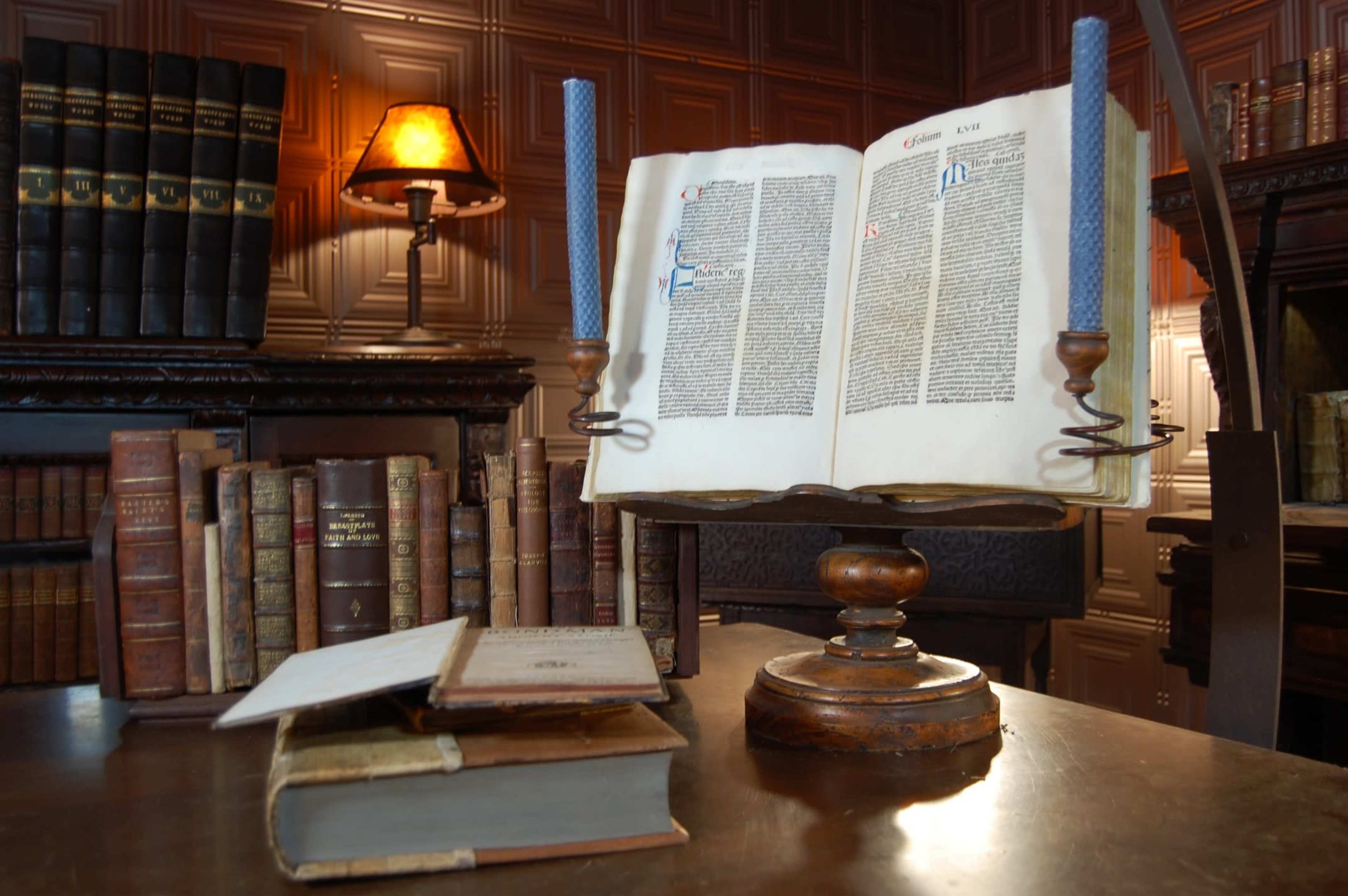 An open book is displayed on a wooden pedestal with two blue candles, surrounded by stacks of vintage books and a warm-lit lamp in the background.