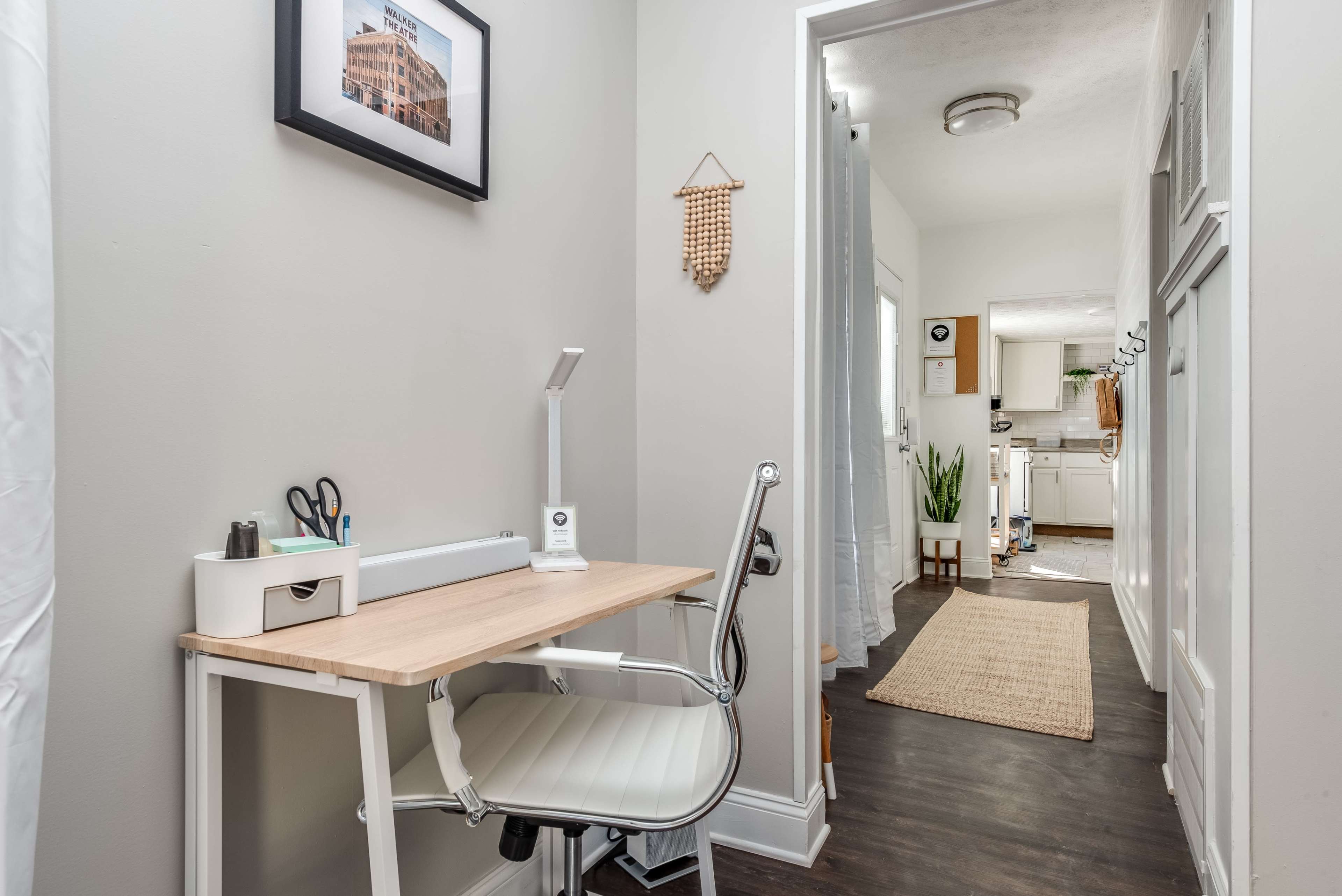 A bright hallway featuring a small workstation with a white chair and wooden desk, leading to a well-lit kitchen area.
