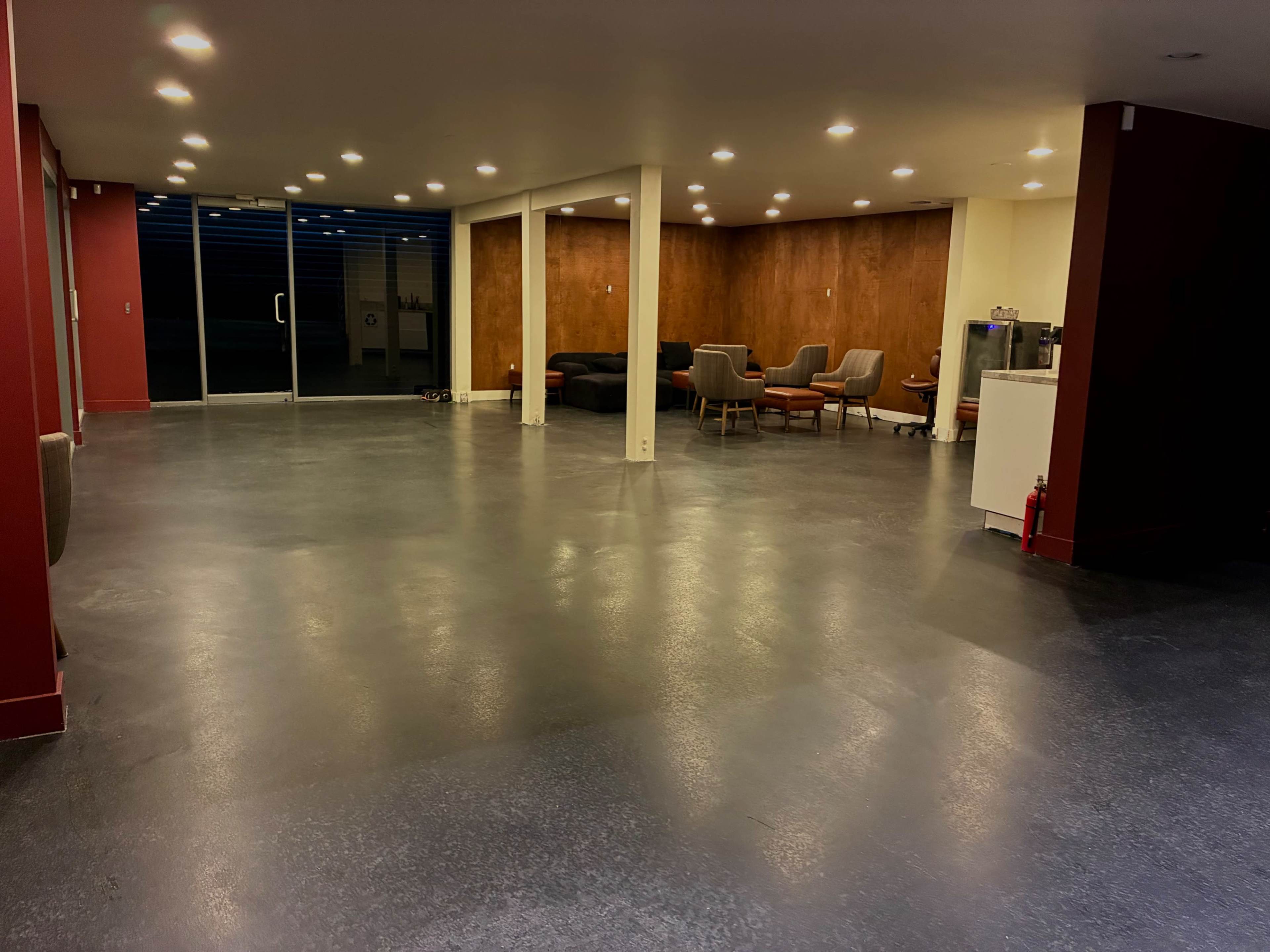 The image shows a spacious, well-lit room with polished concrete flooring, contrasting red walls, and modern seating arranged in a lounge area.