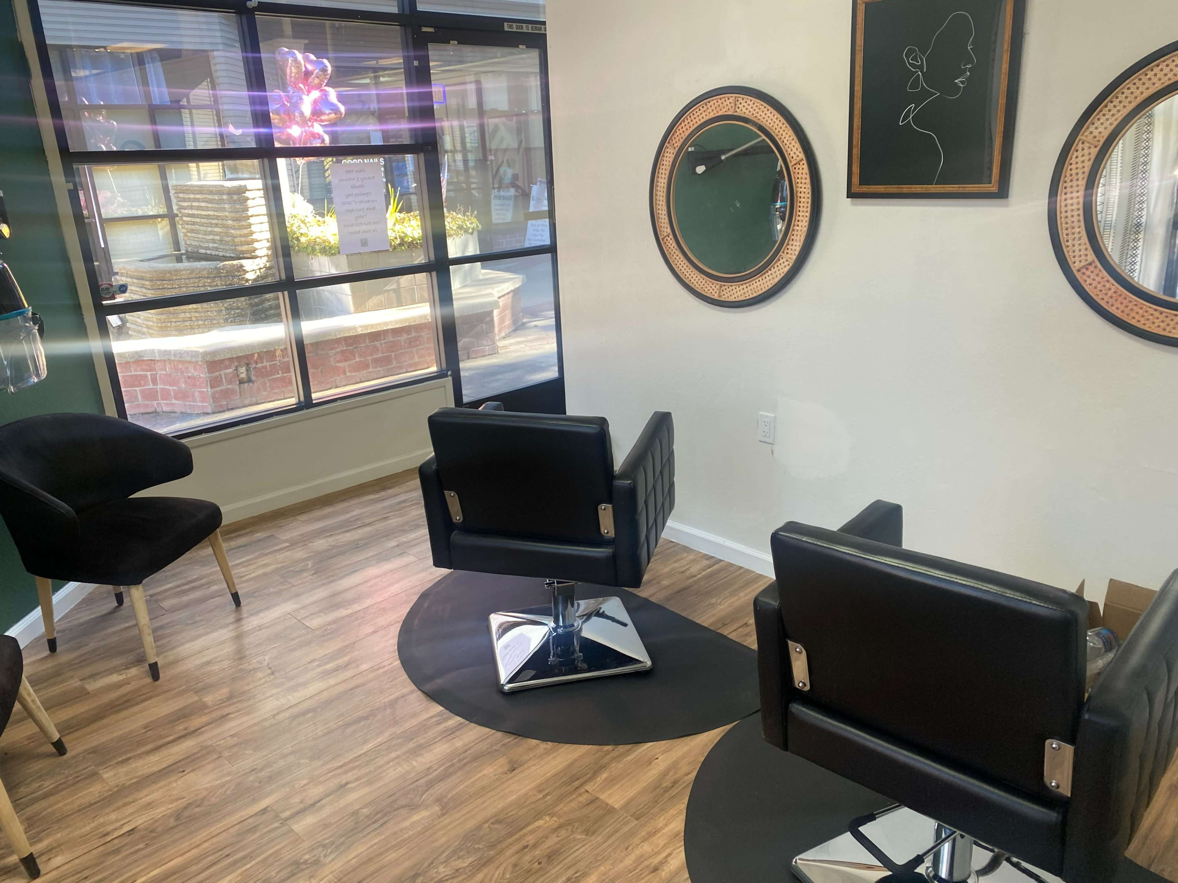A modern hair salon interior featuring two black salon chairs positioned on a circular rug, with mirrors mounted on the wall and natural light coming through large windows.