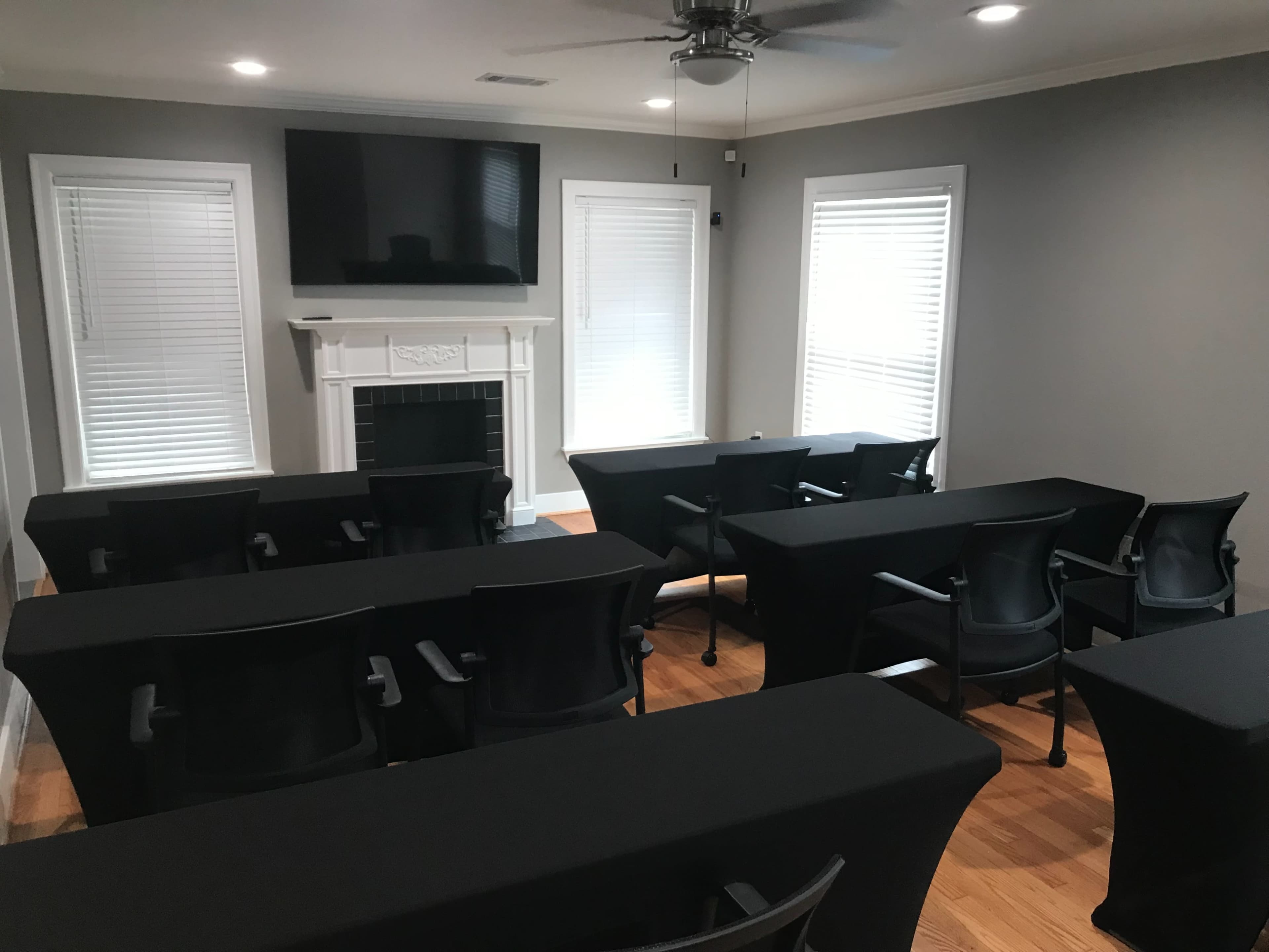 A meeting room featuring several tables with chairs arranged in a classroom style, a television mounted on the wall, and bright windows allowing natural light.