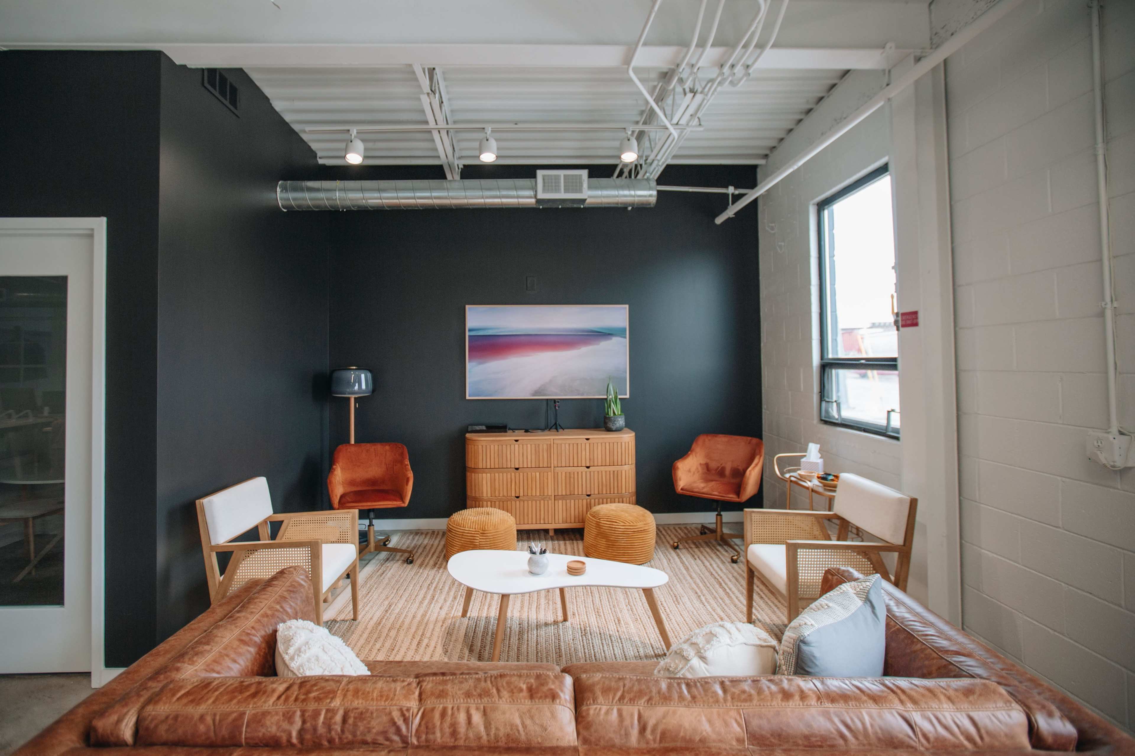 A contemporary office lounge featuring a brown leather sofa, wooden chairs, a white coffee table, and a large wall artwork above a modern wooden cabinet, all set against a dark wall.