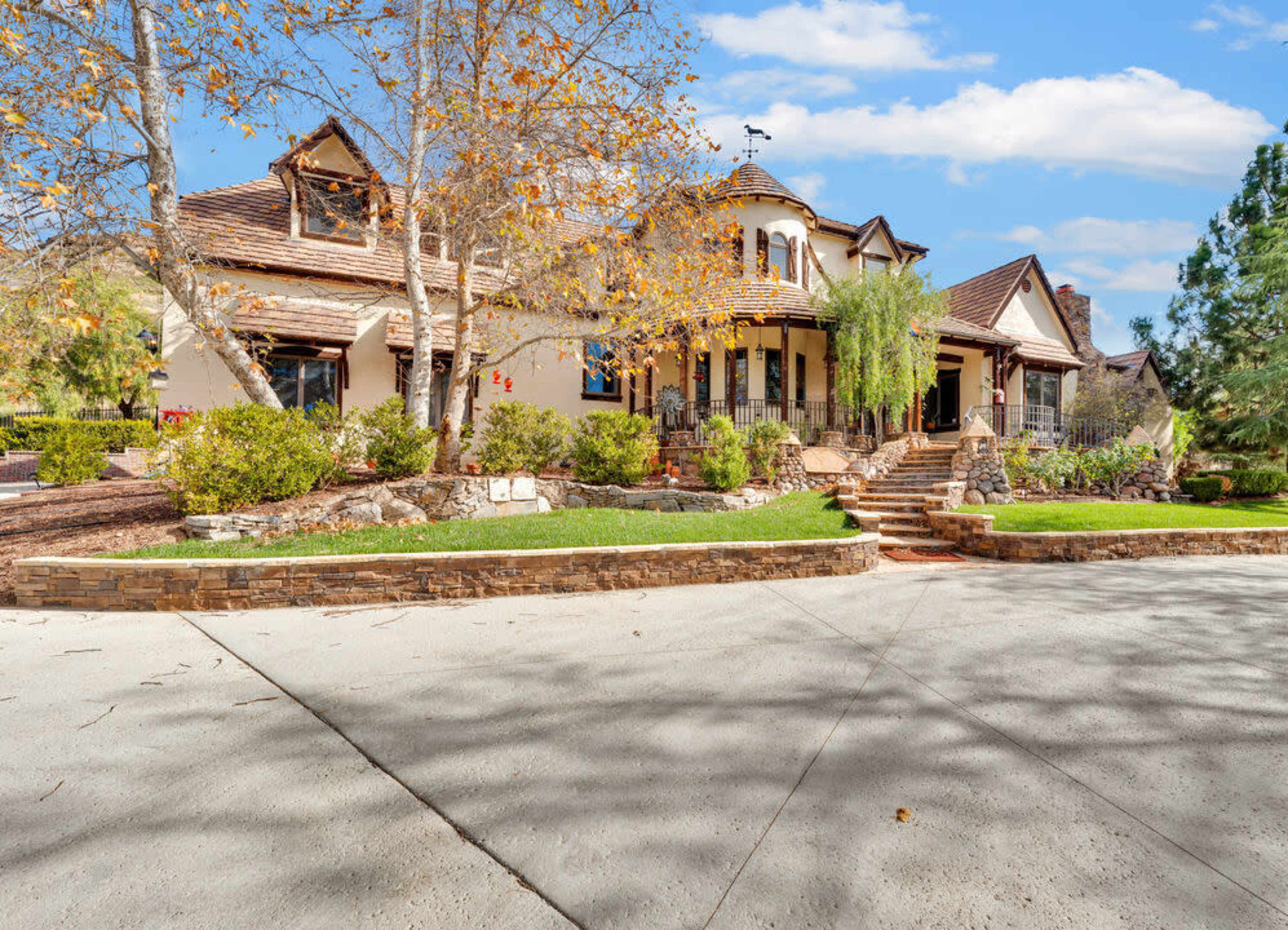A large, multilevel house with a tiled roof and various shrubs, surrounded by a paved driveway and trees.