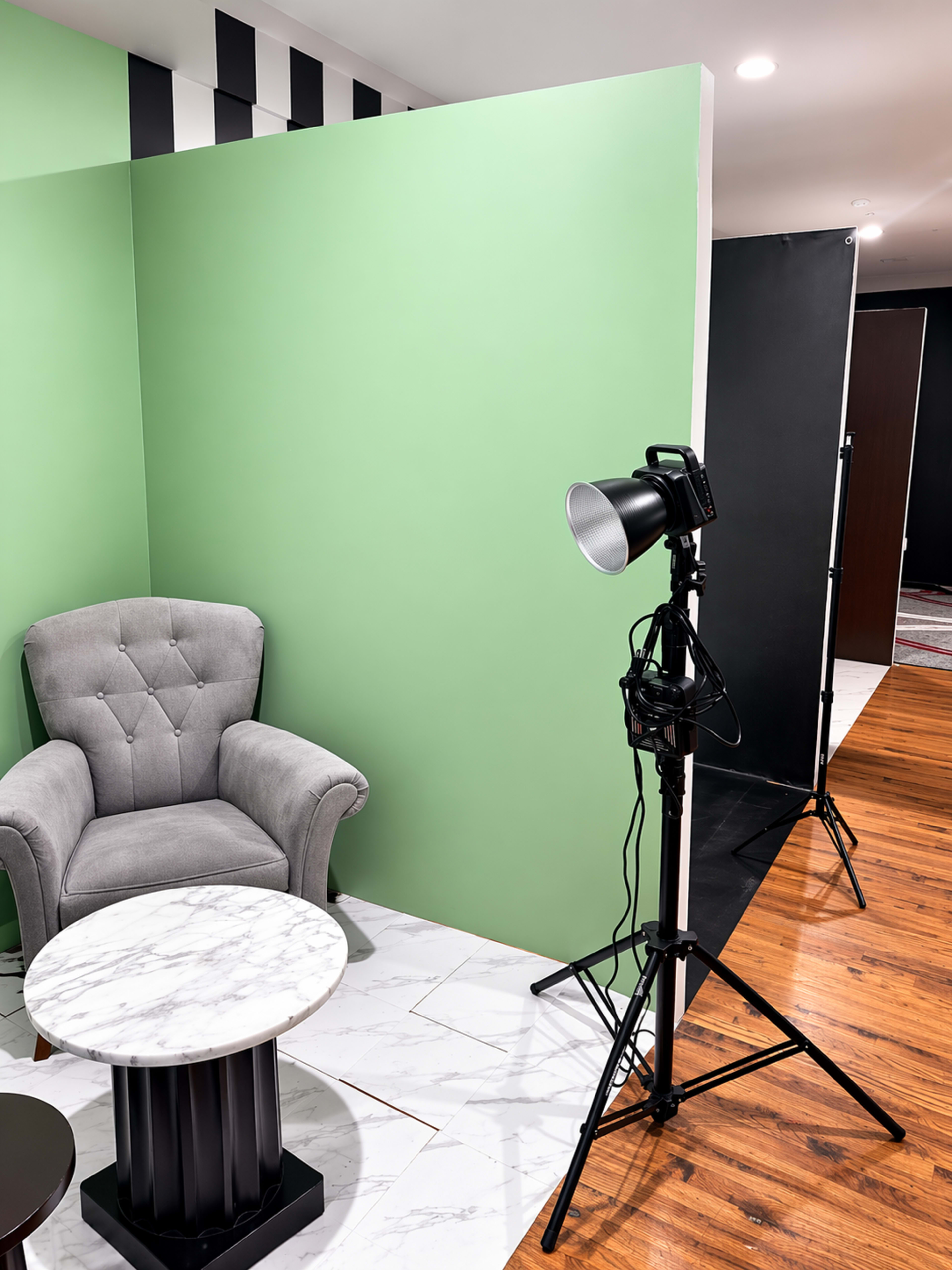 The image shows a gray upholstered armchair next to a round marble table, illuminated by a spotlight, against a green wall in a room with contrasting black and white elements.