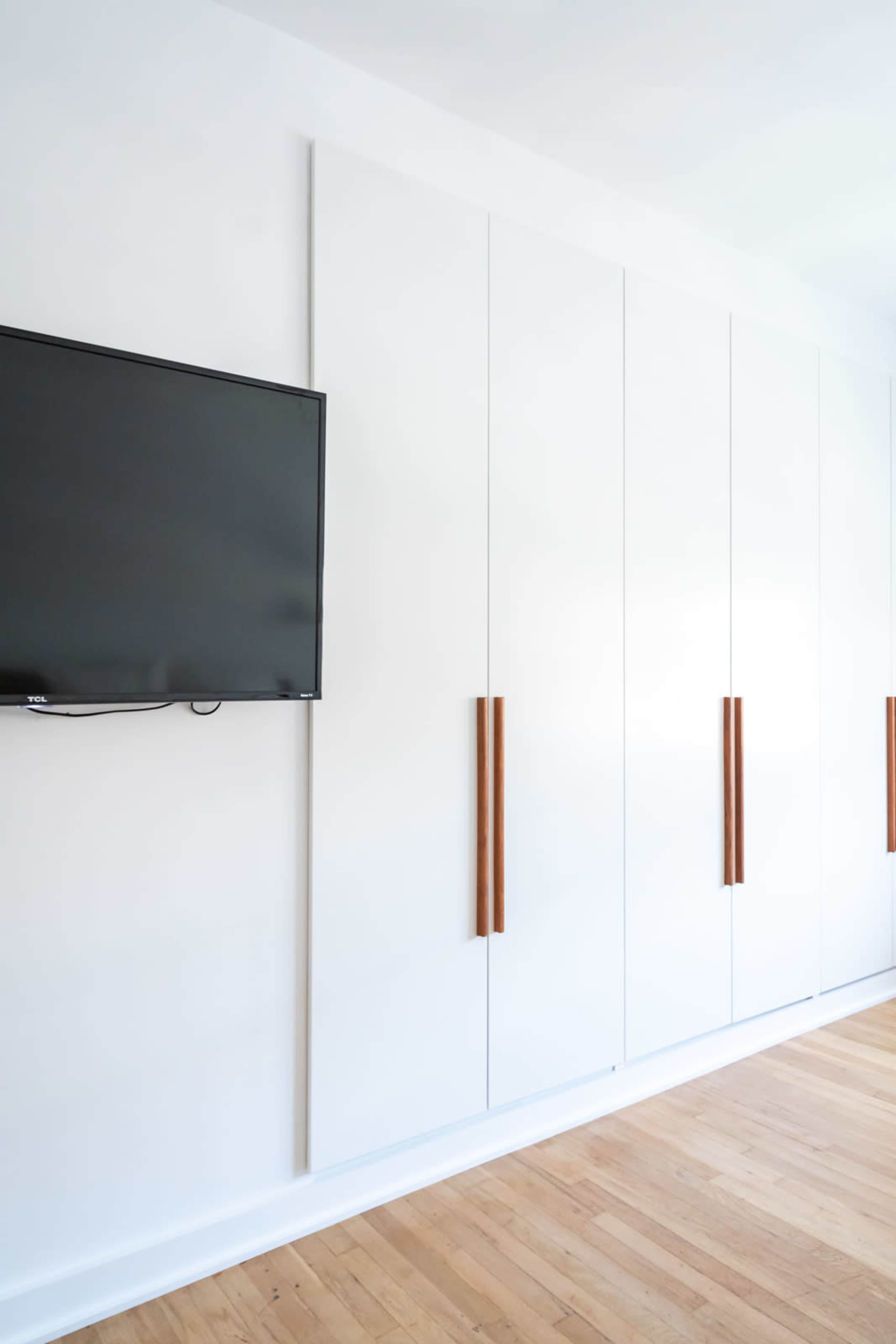 A flat-screen television is mounted on a white wall next to a large white cabinet with wooden handles.