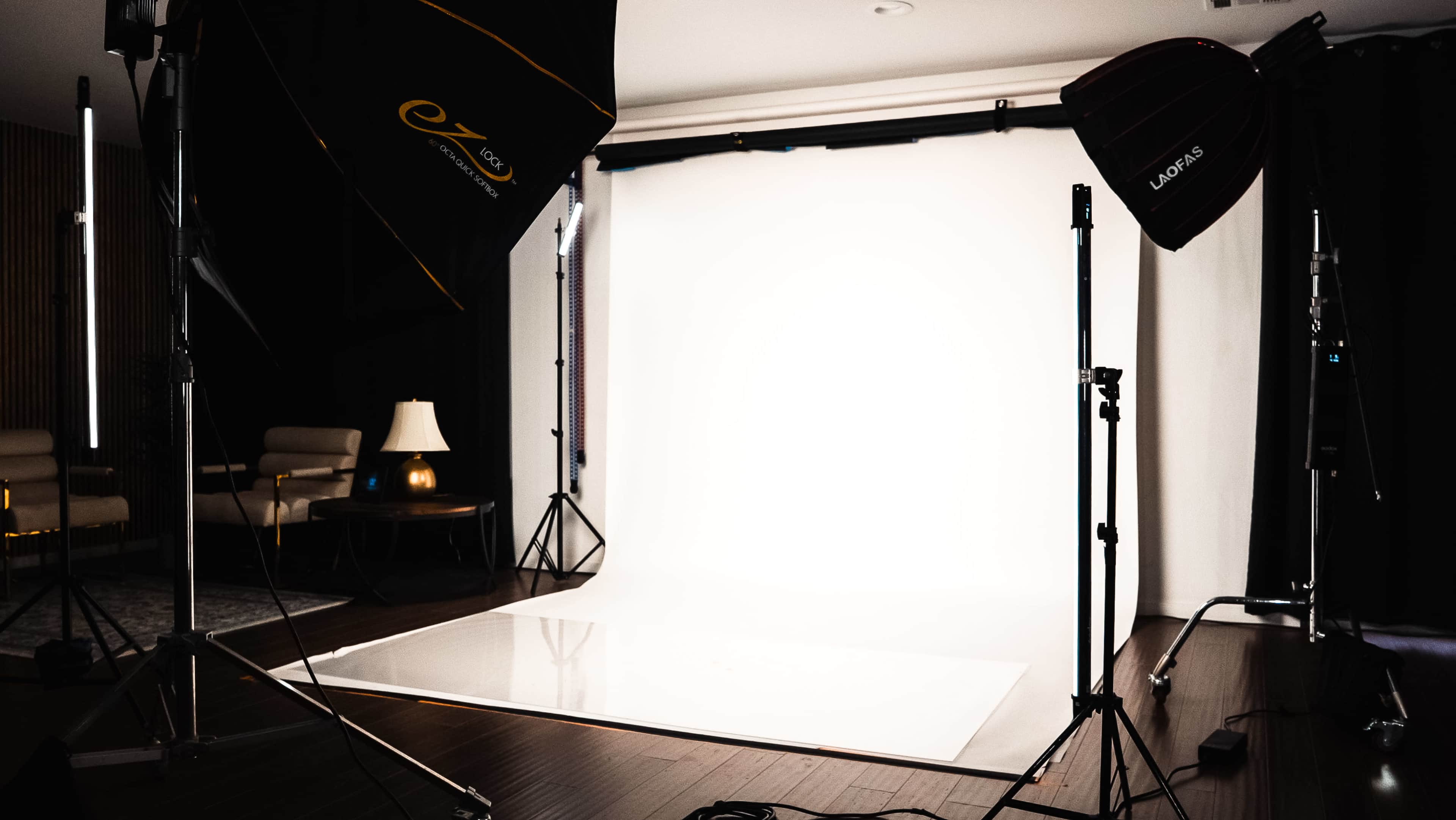 The image shows a photography studio setup with a white backdrop, professional lighting equipment, and minimal furniture.