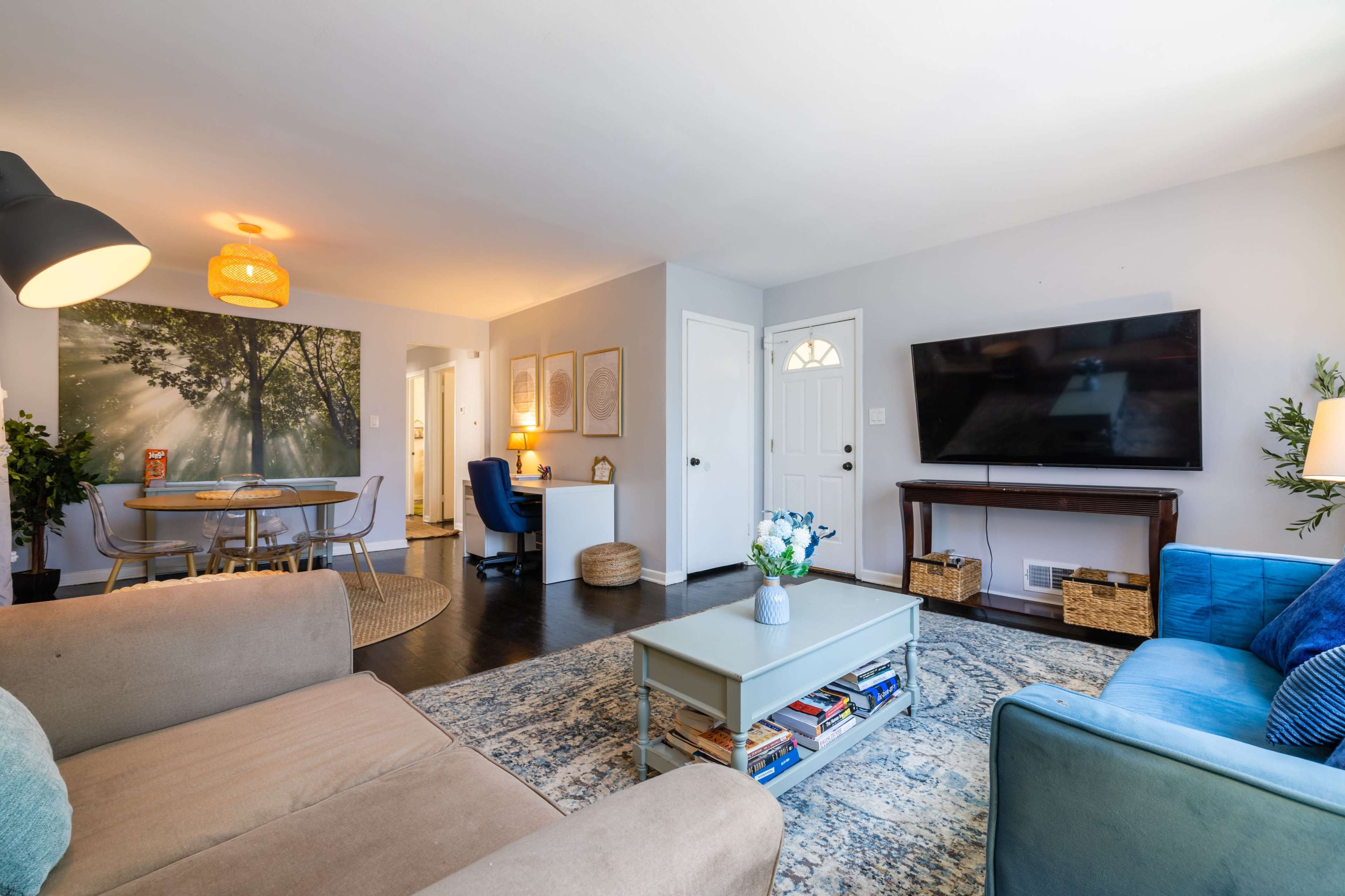 The image shows a modern living room with a blue couch, a tan sofa, a coffee table, and a large television, featuring a dining area and workspace in the background.