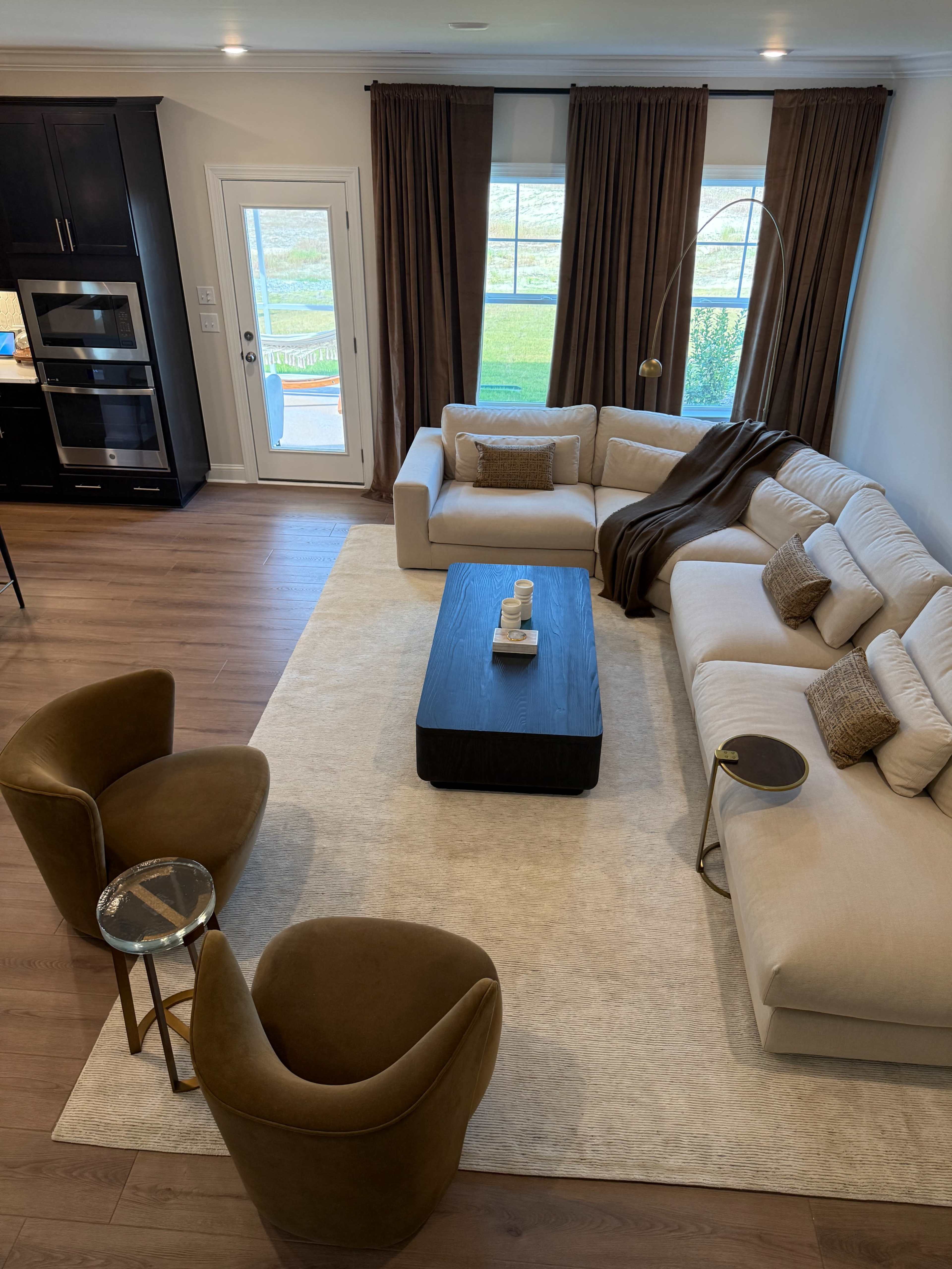 The image shows a modern living room featuring a large sectional sofa, two brown chairs, a coffee table, and large windows with curtains.