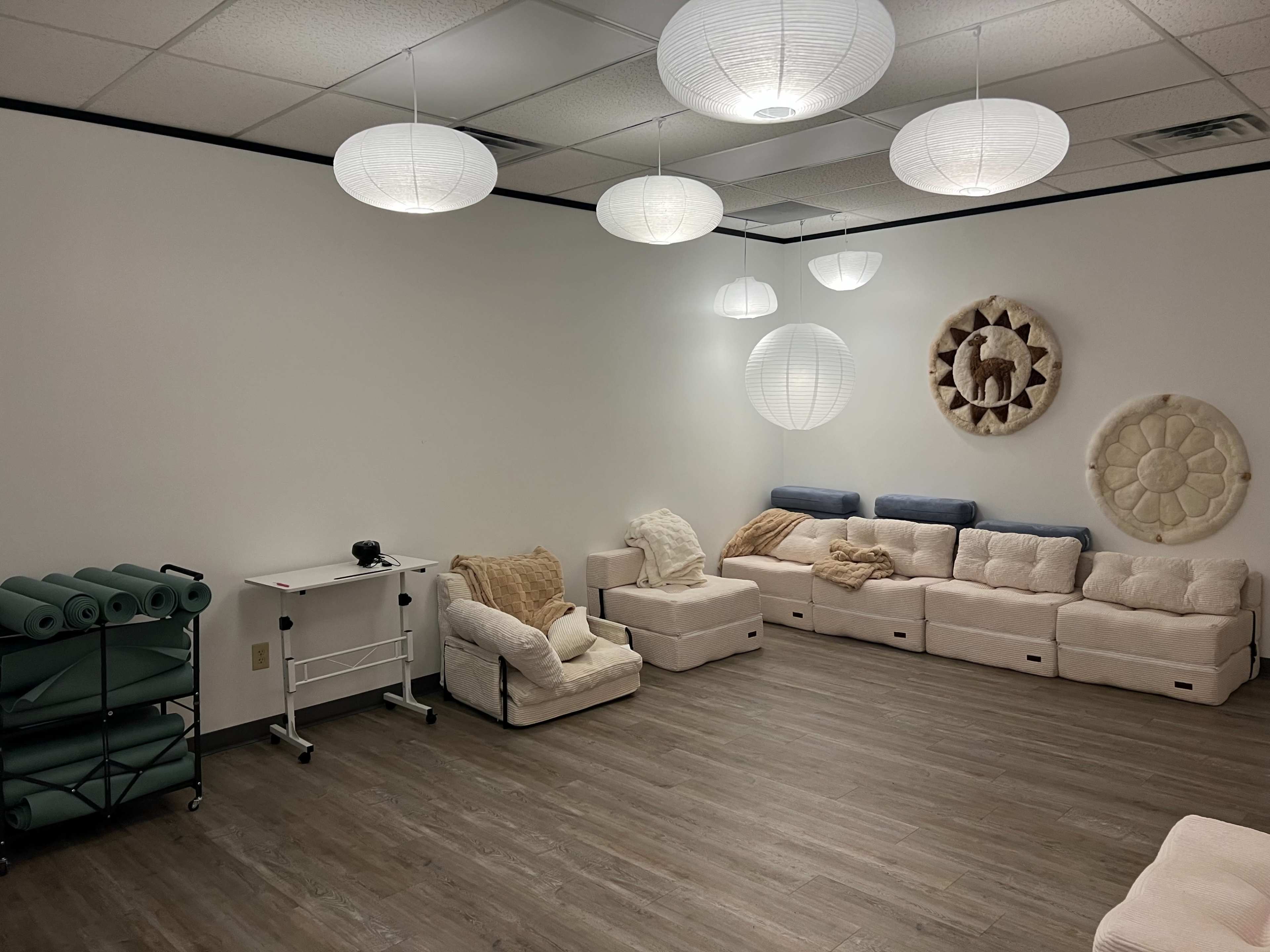 The image shows a spacious, minimalist room with light-colored seating, yoga mats stacked in a corner, and paper lanterns hanging from the ceiling.