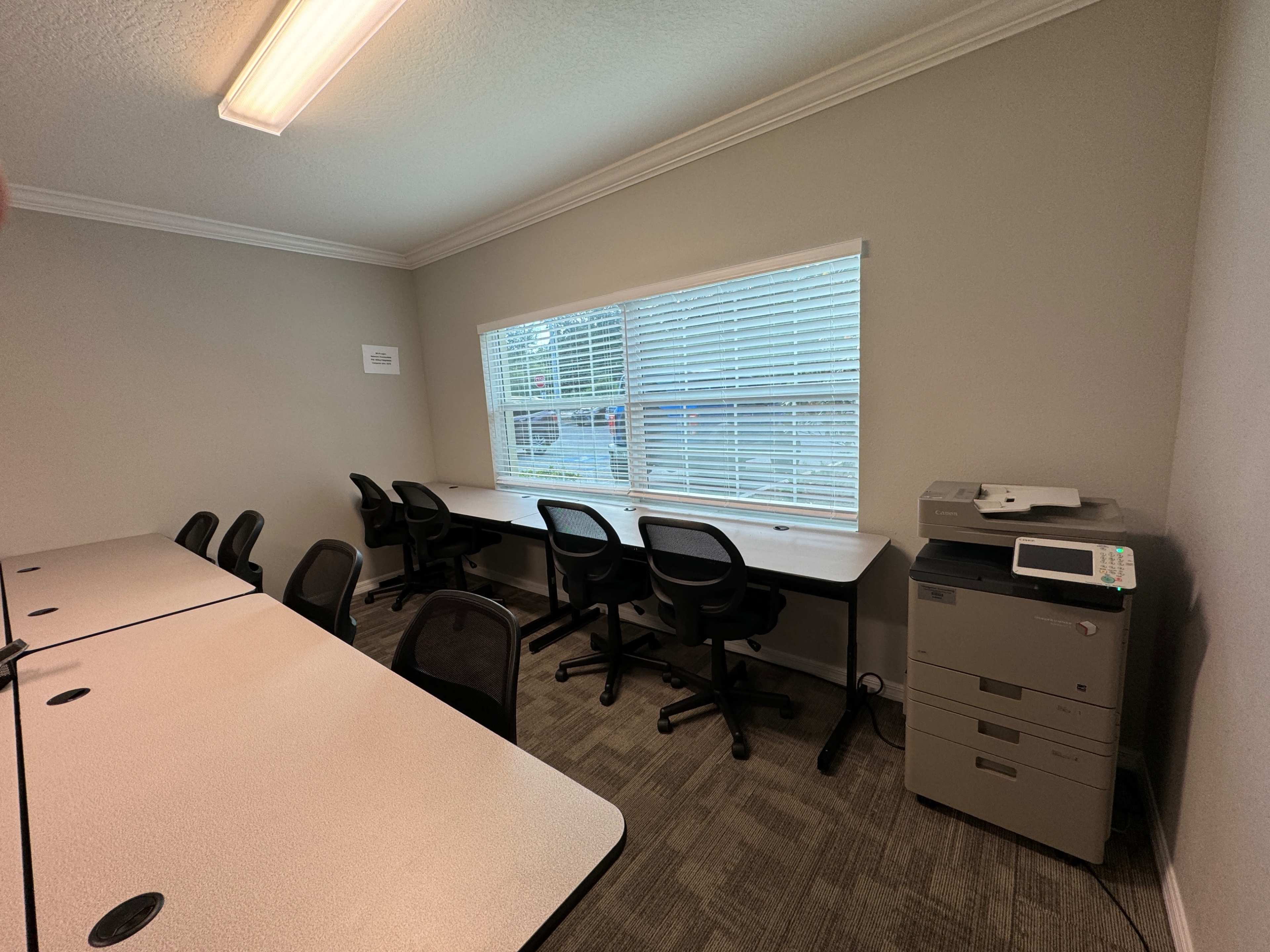 A small office space with a row of chairs at a long table, a window with blinds, and a printer next to it.