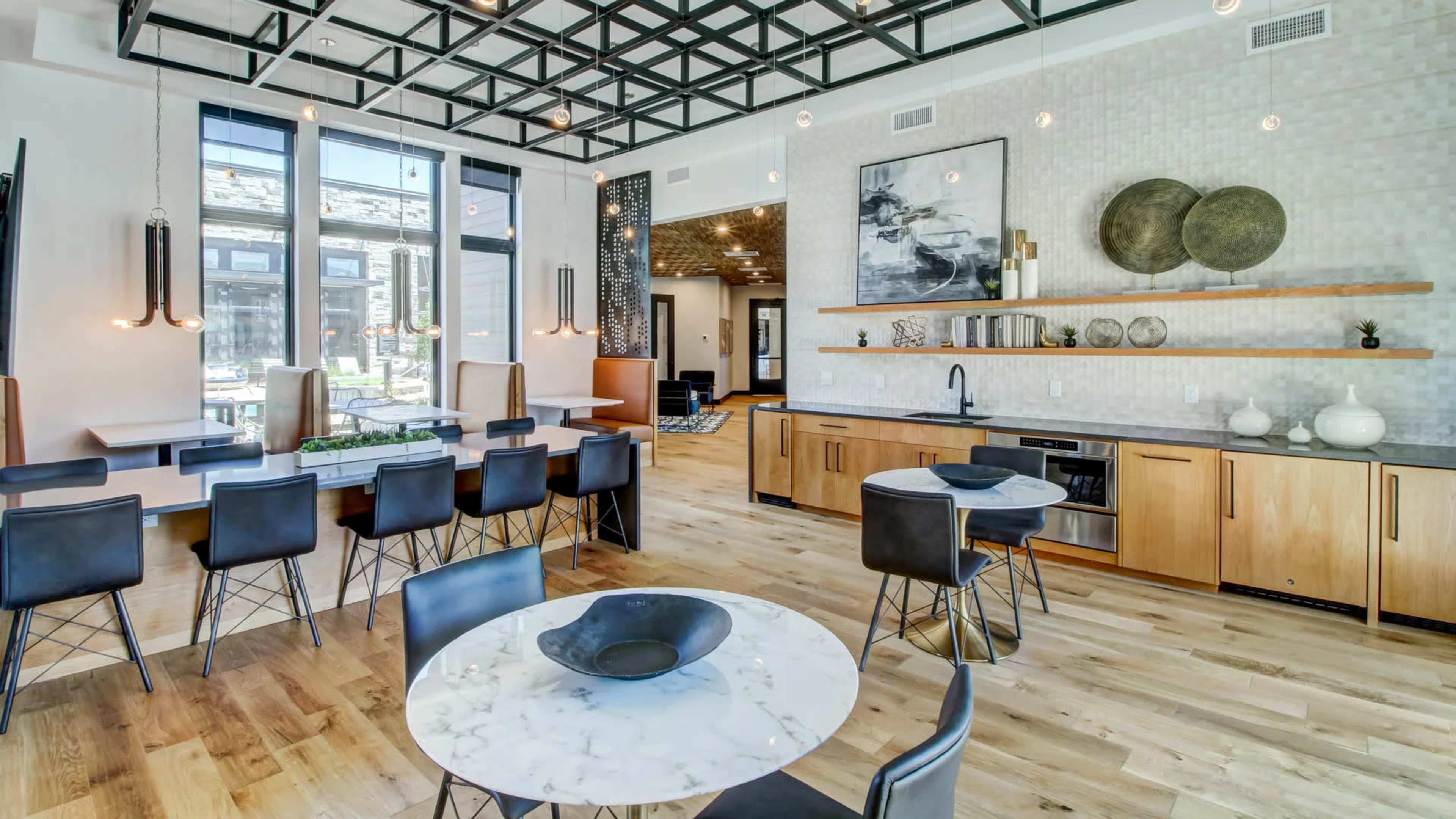 A modern kitchen and dining area features a large tabletop, sleek black chairs, wooden cabinetry, and decorative items against a textured wall.