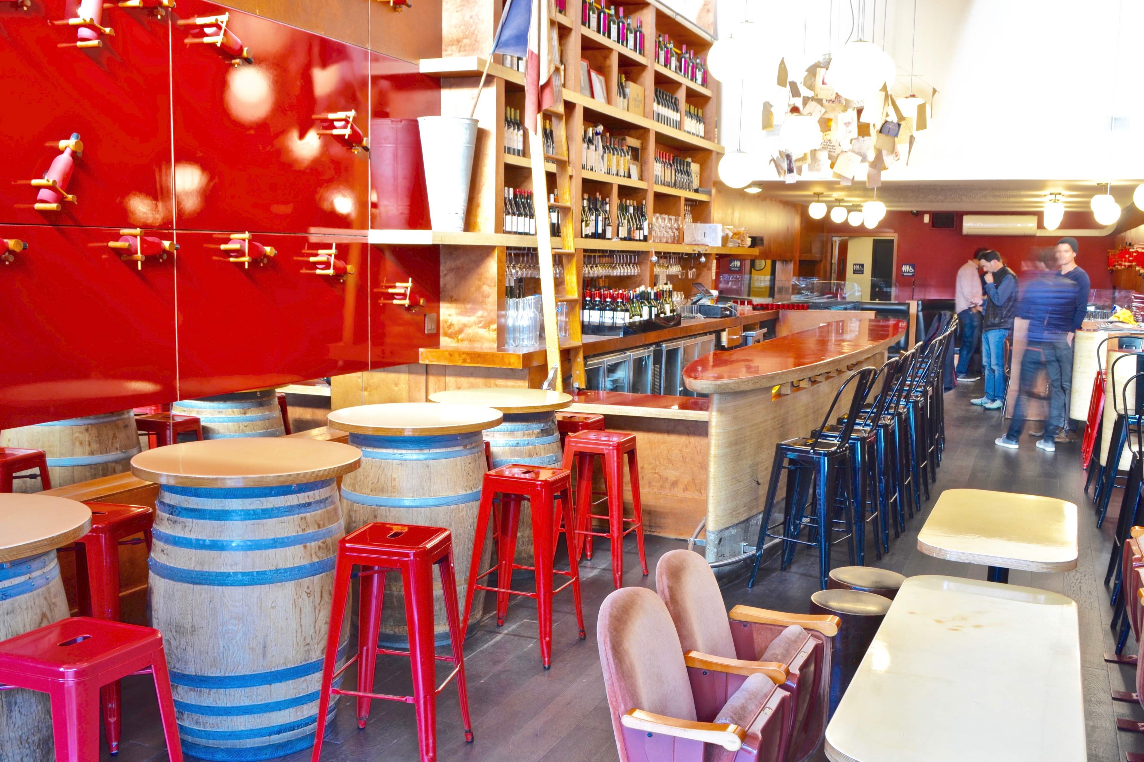 A bar interior with red walls, wooden shelves lined with bottles, and a mix of barrel and wooden seating.