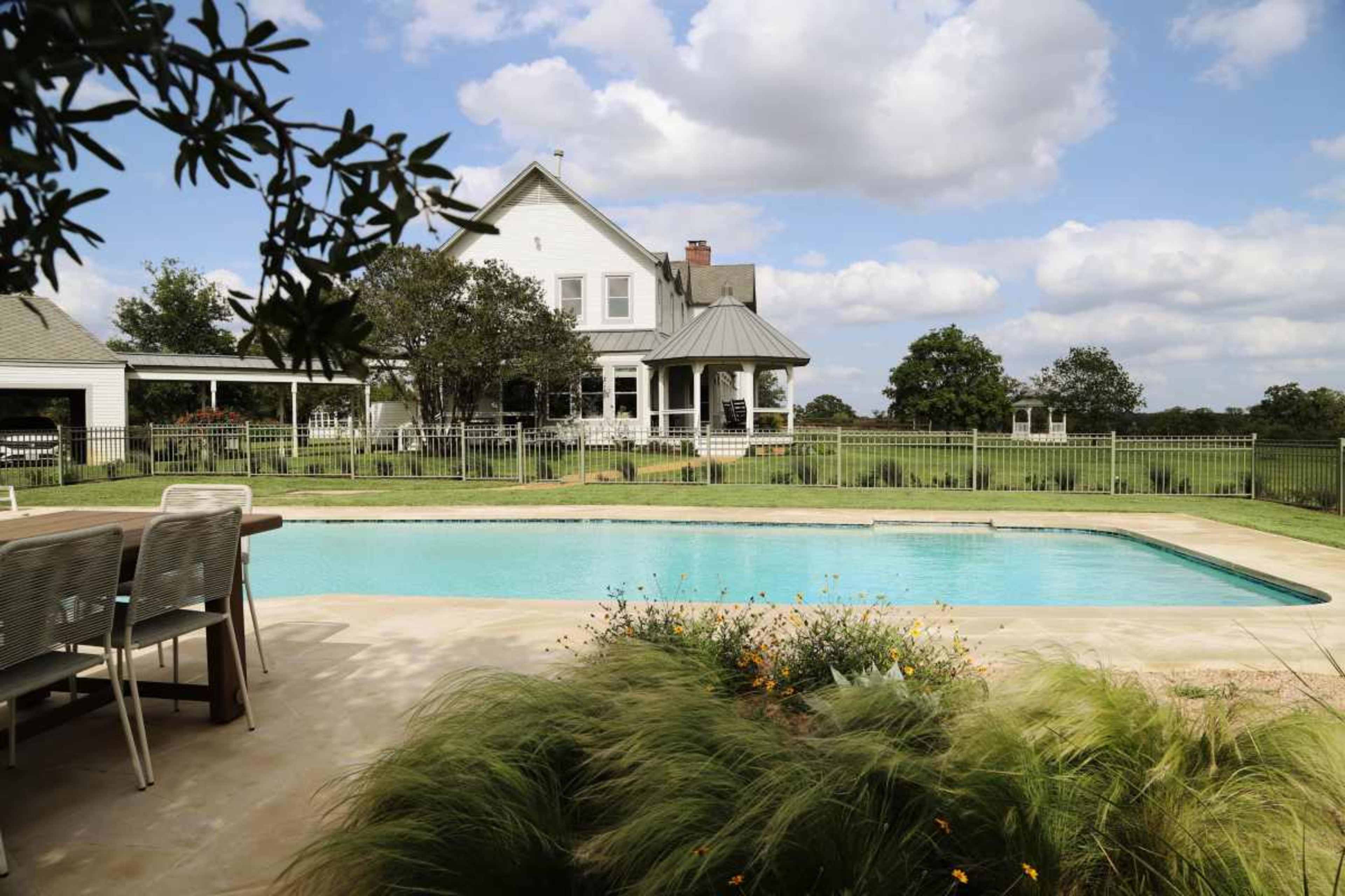 The image shows a spacious backyard featuring a swimming pool, a dining area, and a large white house with a porch set against a backdrop of blue skies and greenery.