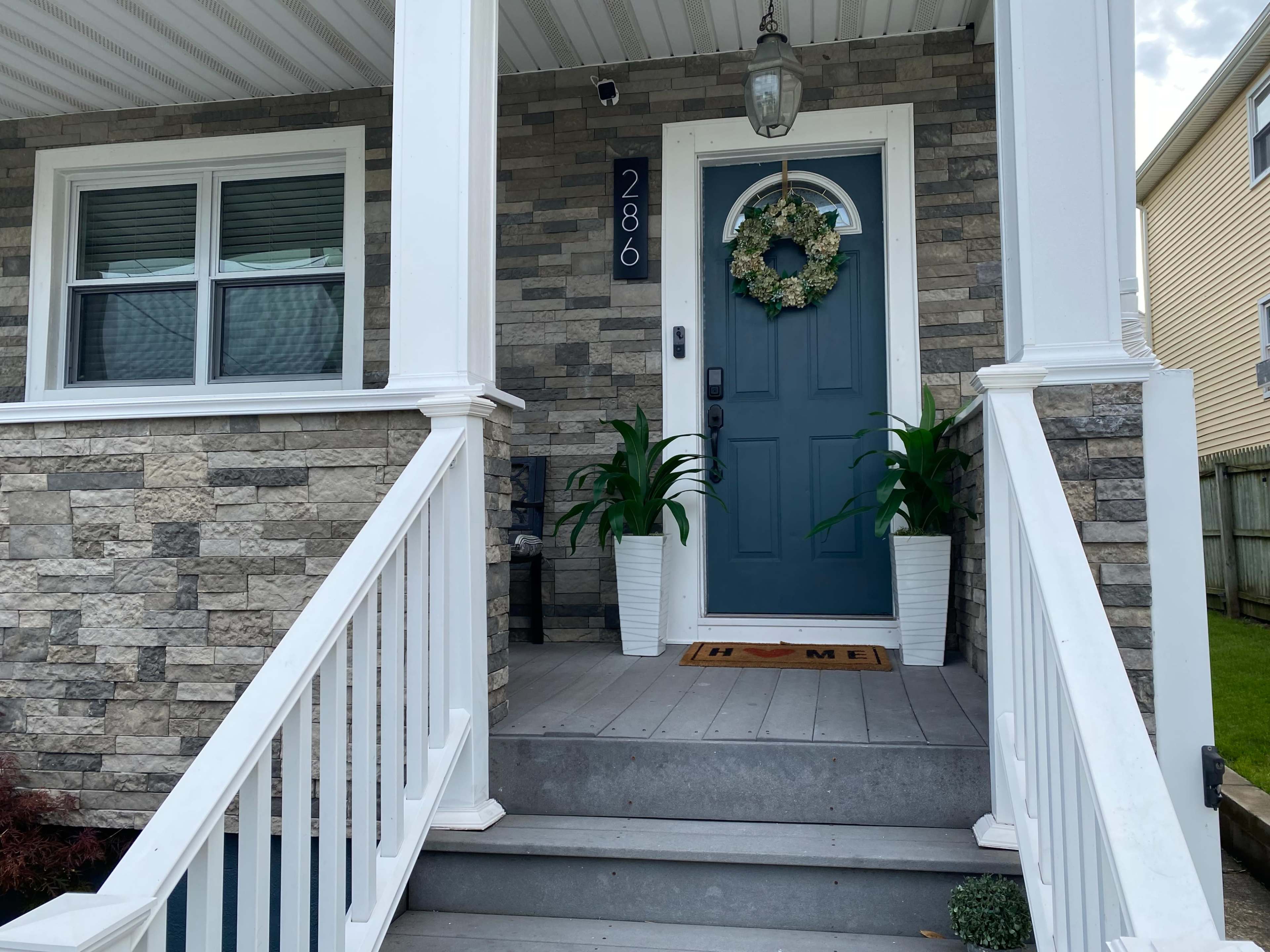 The image shows a front entrance with a dark blue door, flanked by two potted plants, a welcome mat, and a stone wall facade.