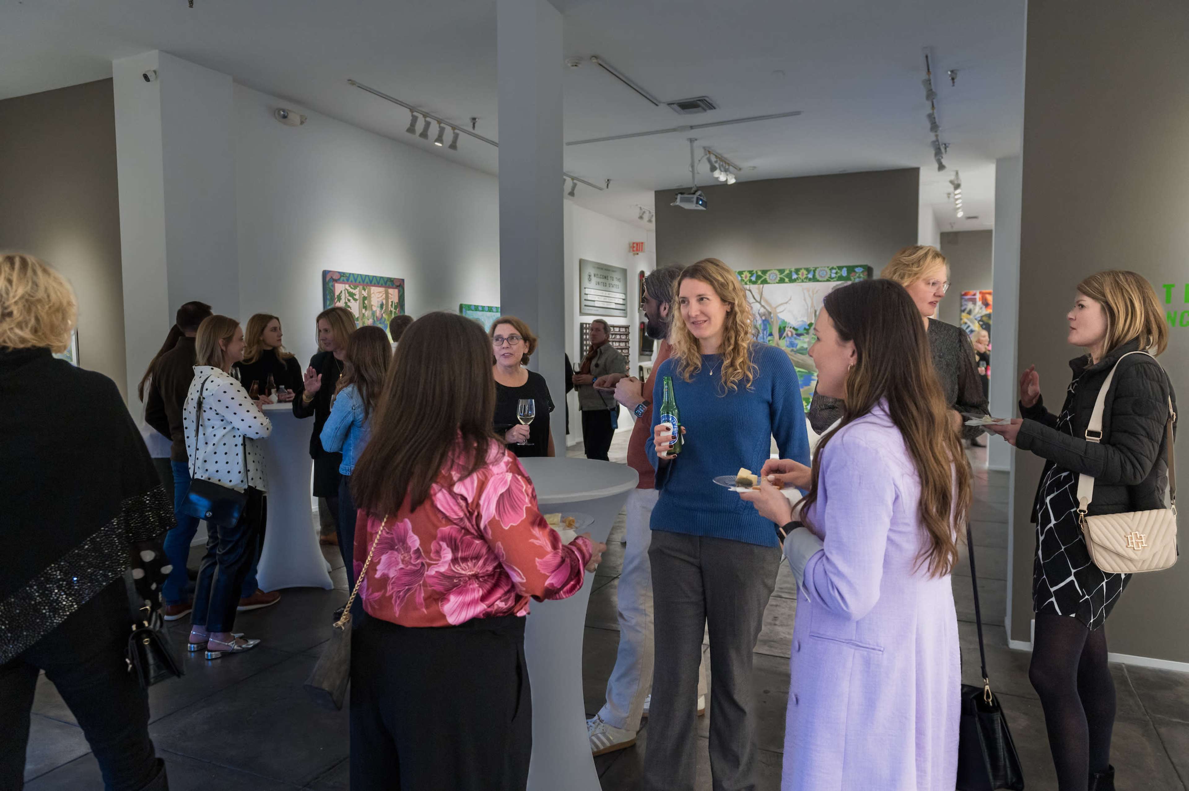 A group of people is socializing at an art gallery event, holding drinks and conversation near white display tables.