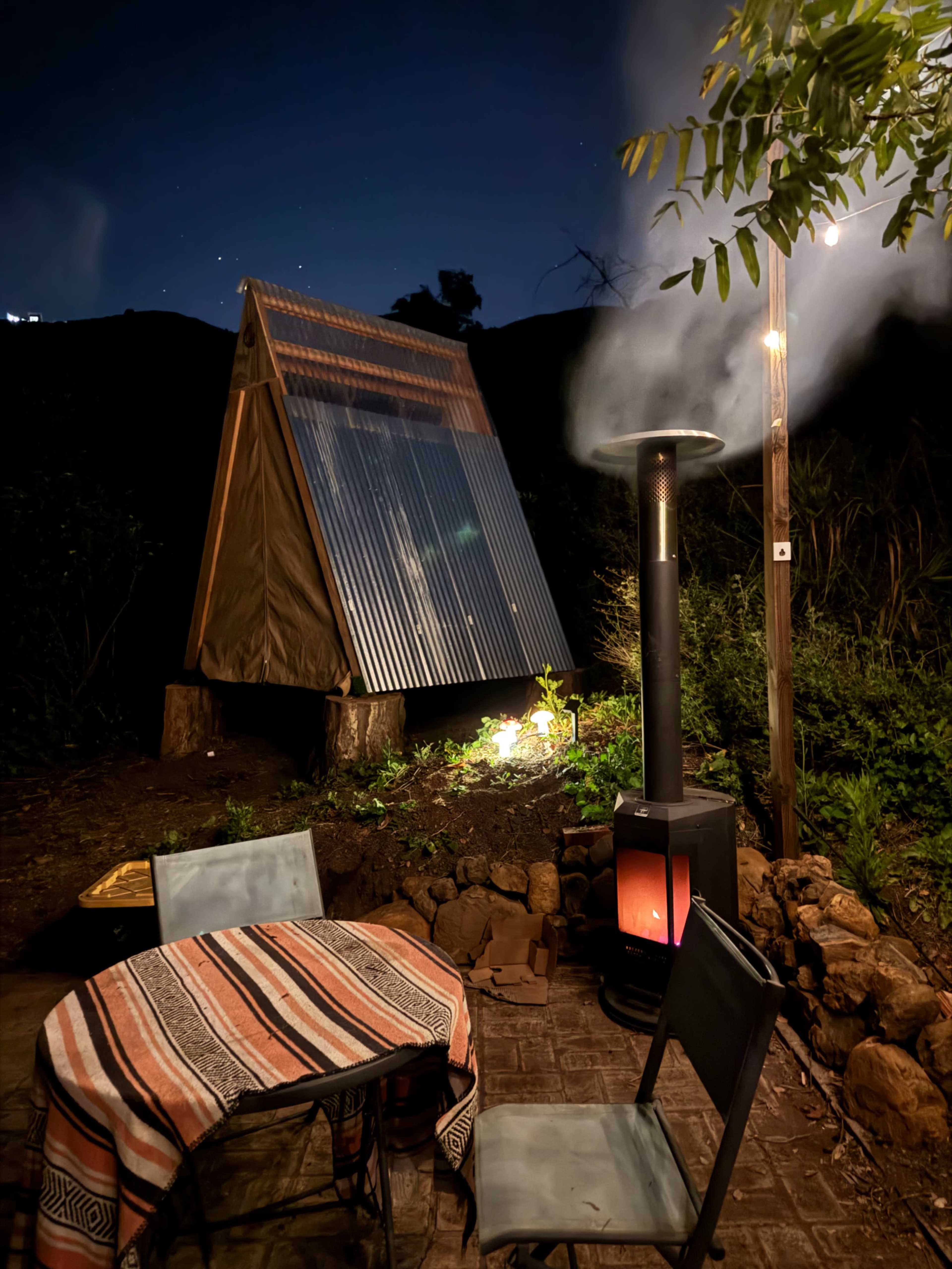 A small tent-like structure with a corrugated metal roof is illuminated by a nearby light, alongside a table and chairs near a patio heater, all set in a garden at night.