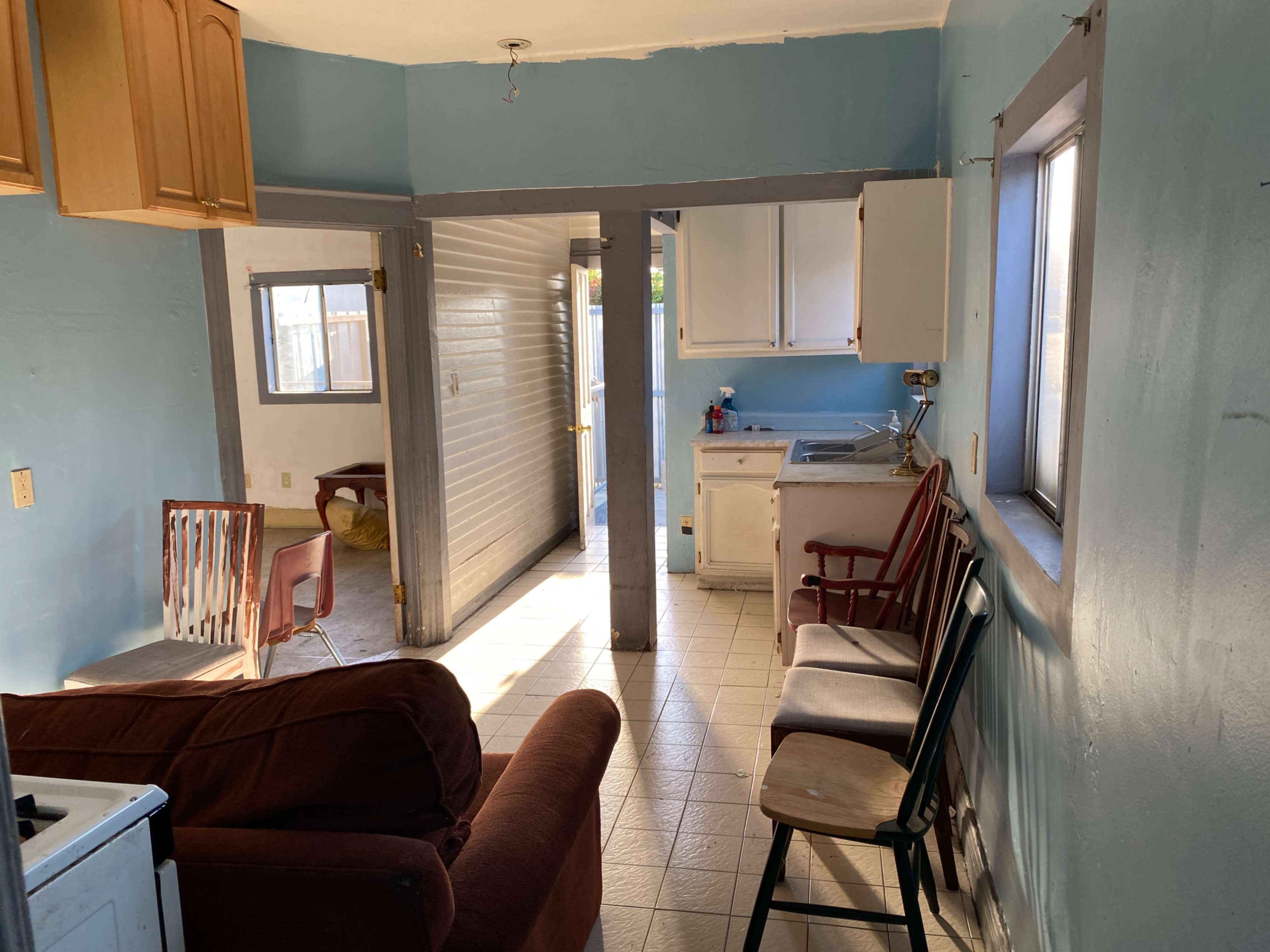 A small, light blue living area connected to a kitchen with a sofa, two chairs, and bright sunlight entering through the windows.