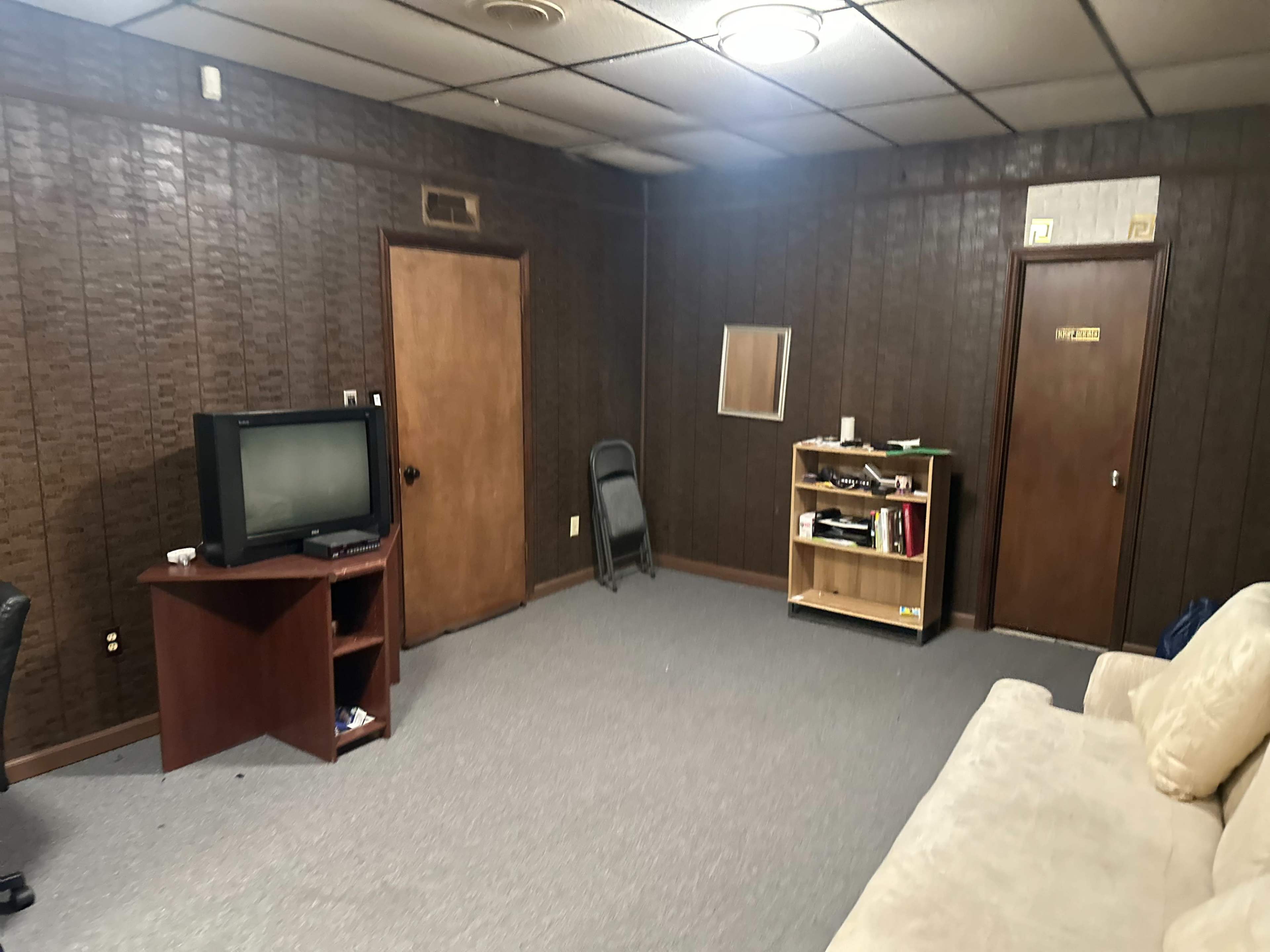The image shows a sparsely furnished room with dark wood paneling, featuring a television on a stand, a rolling chair, a bookshelf, and a door.