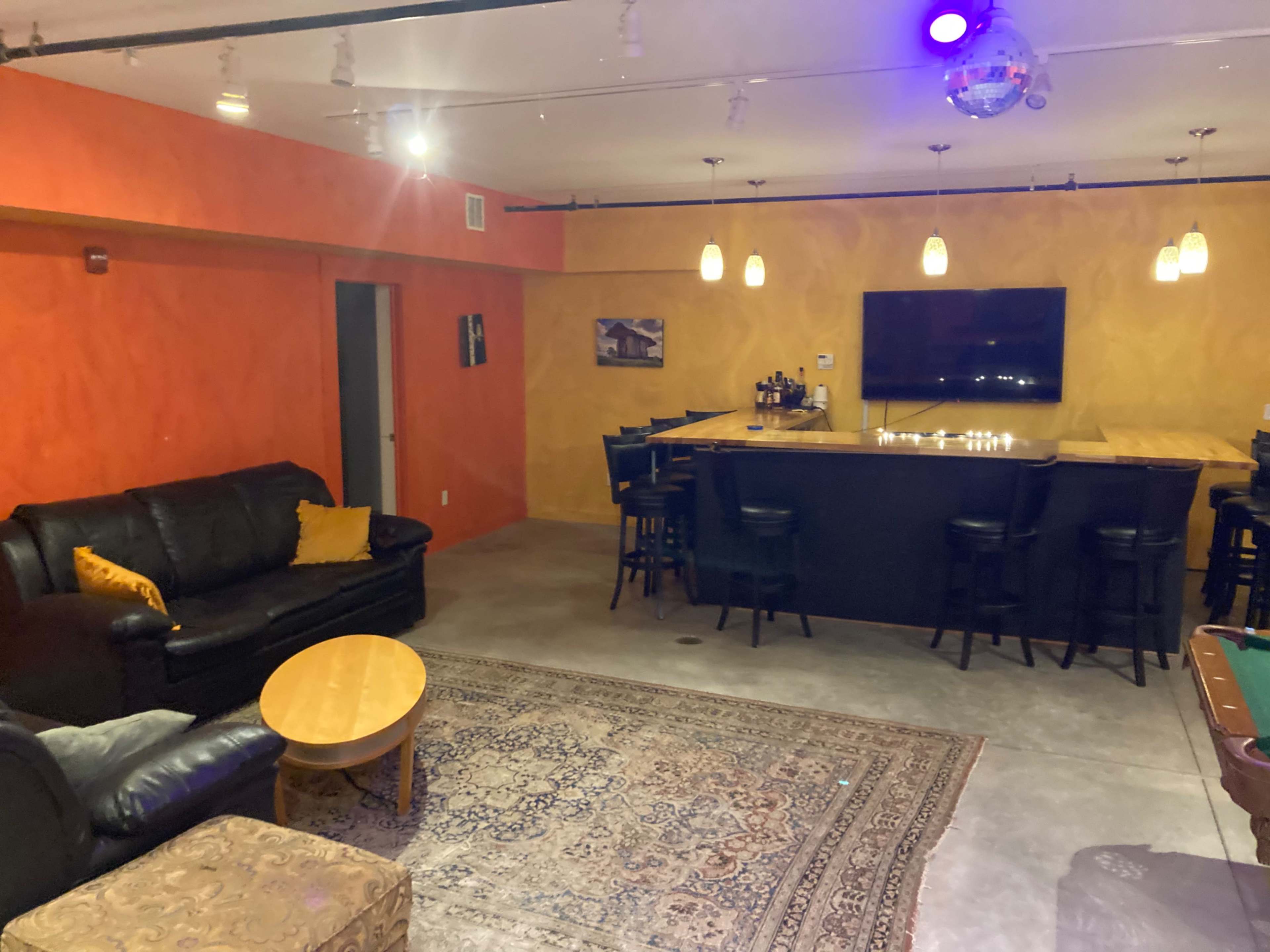 A colorful lounge area featuring a black leather couch, a round wooden coffee table, a bar with high stools, and a pool table, all illuminated by ambient lighting.