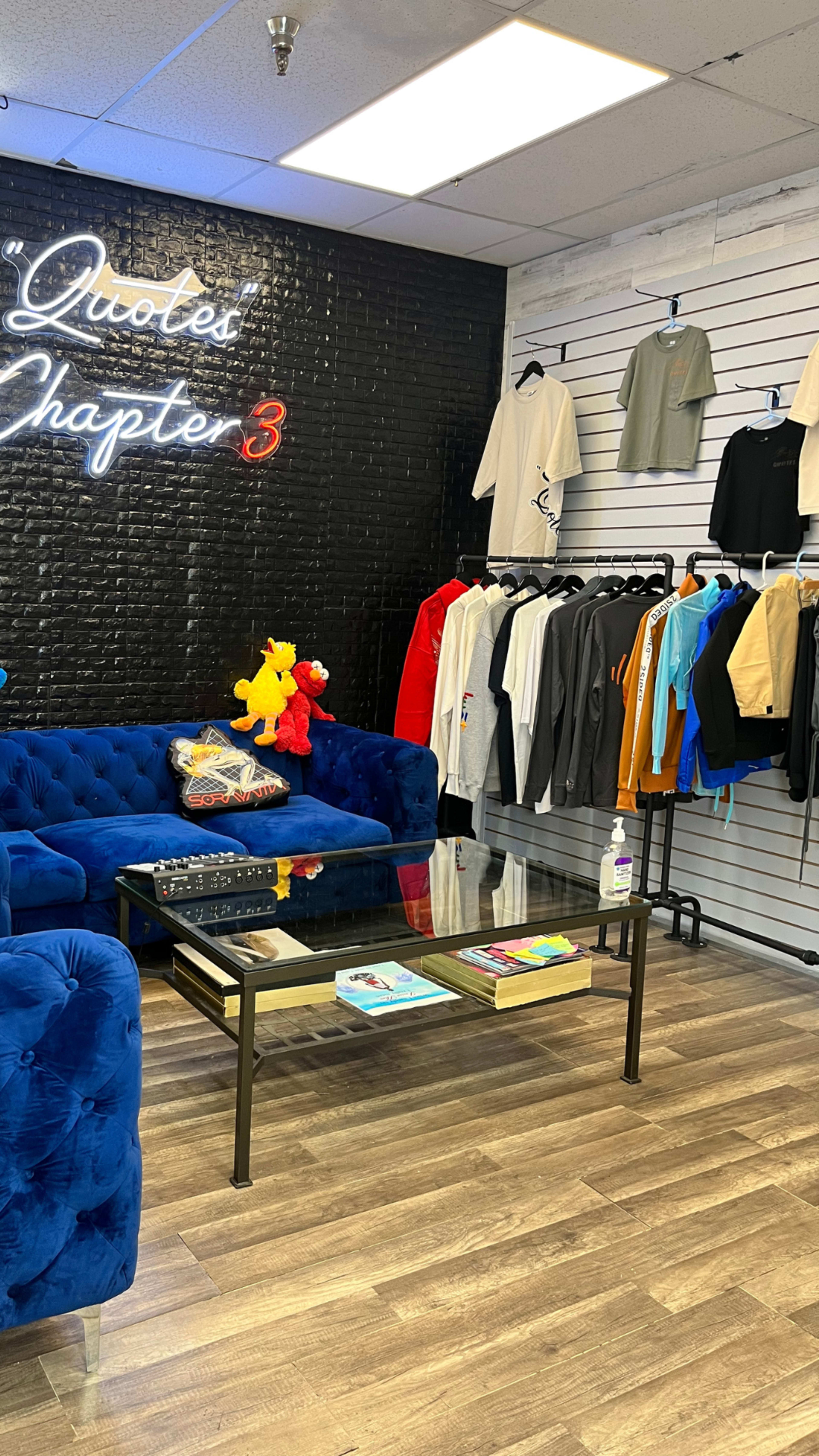 A modern retail space features a blue velvet couch, a glass coffee table, and a display of clothing on hangers against a black wall with illuminated text.