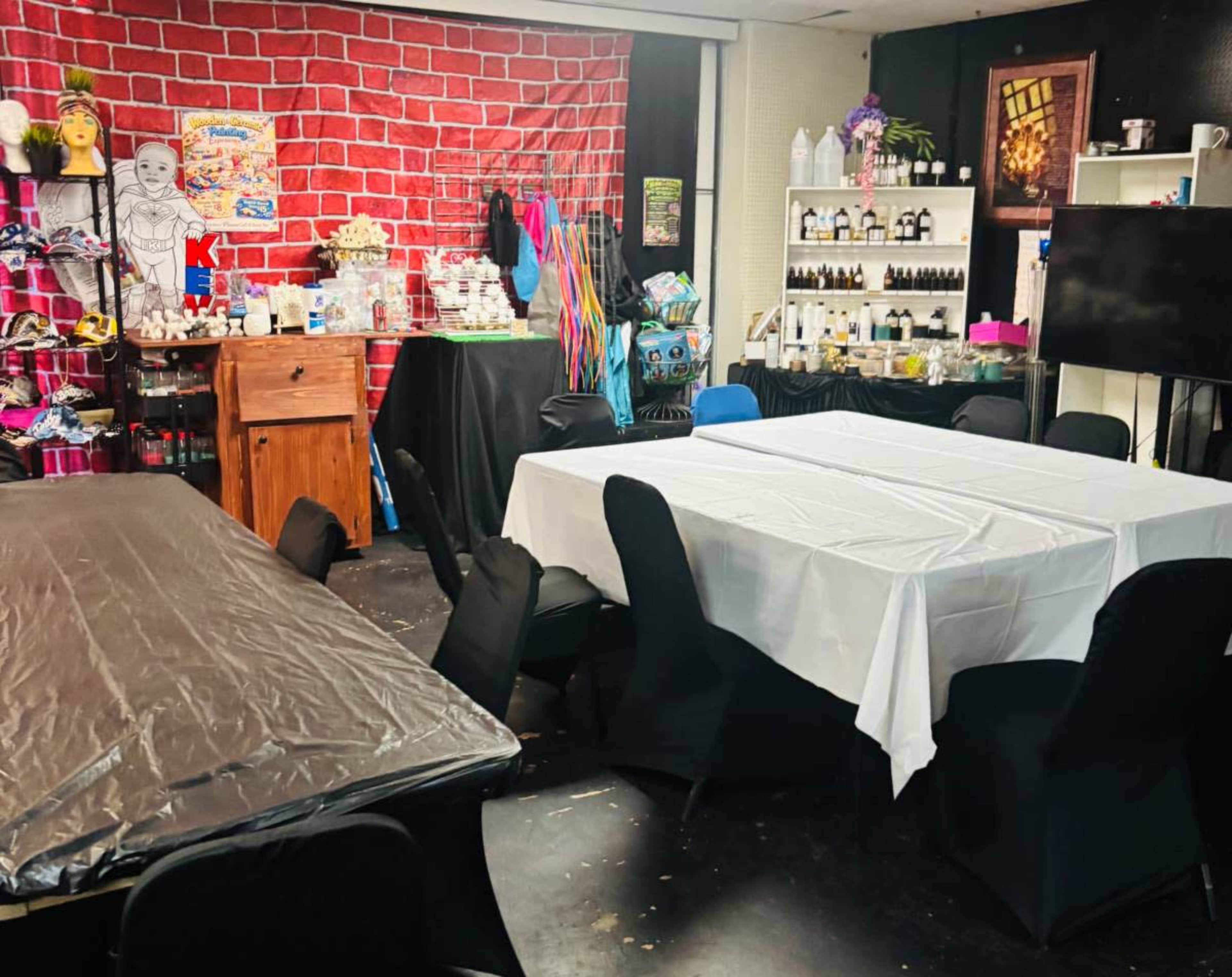 The image shows a room set up for an event, featuring several tables covered with black and white tablecloths, a vibrant red brick wall in the background, and various supplies displayed on shelves.