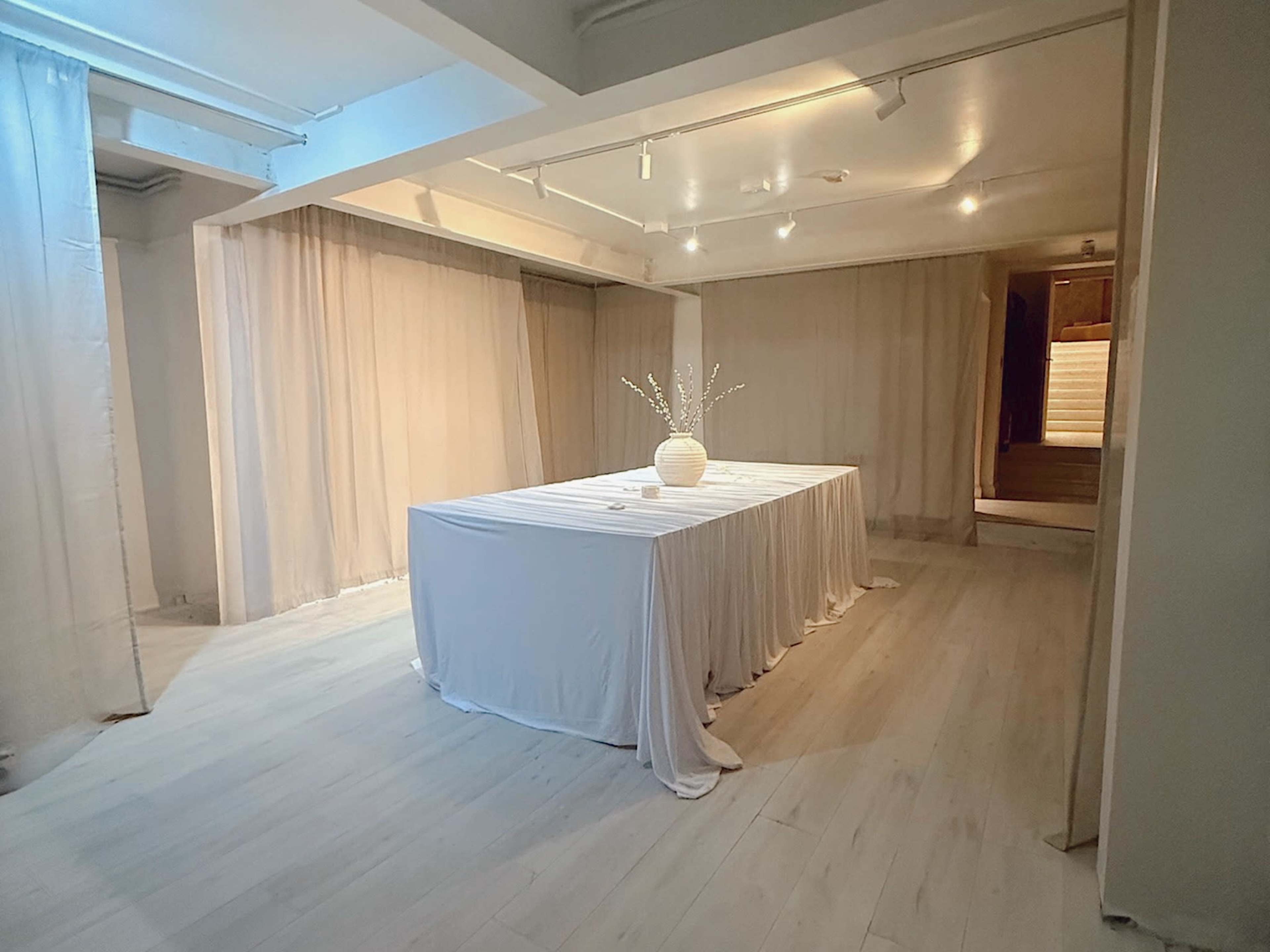 The image shows a spacious room with a long, white-draped table at the center, surrounded by soft fabric curtains and warmly lit by overhead lights.