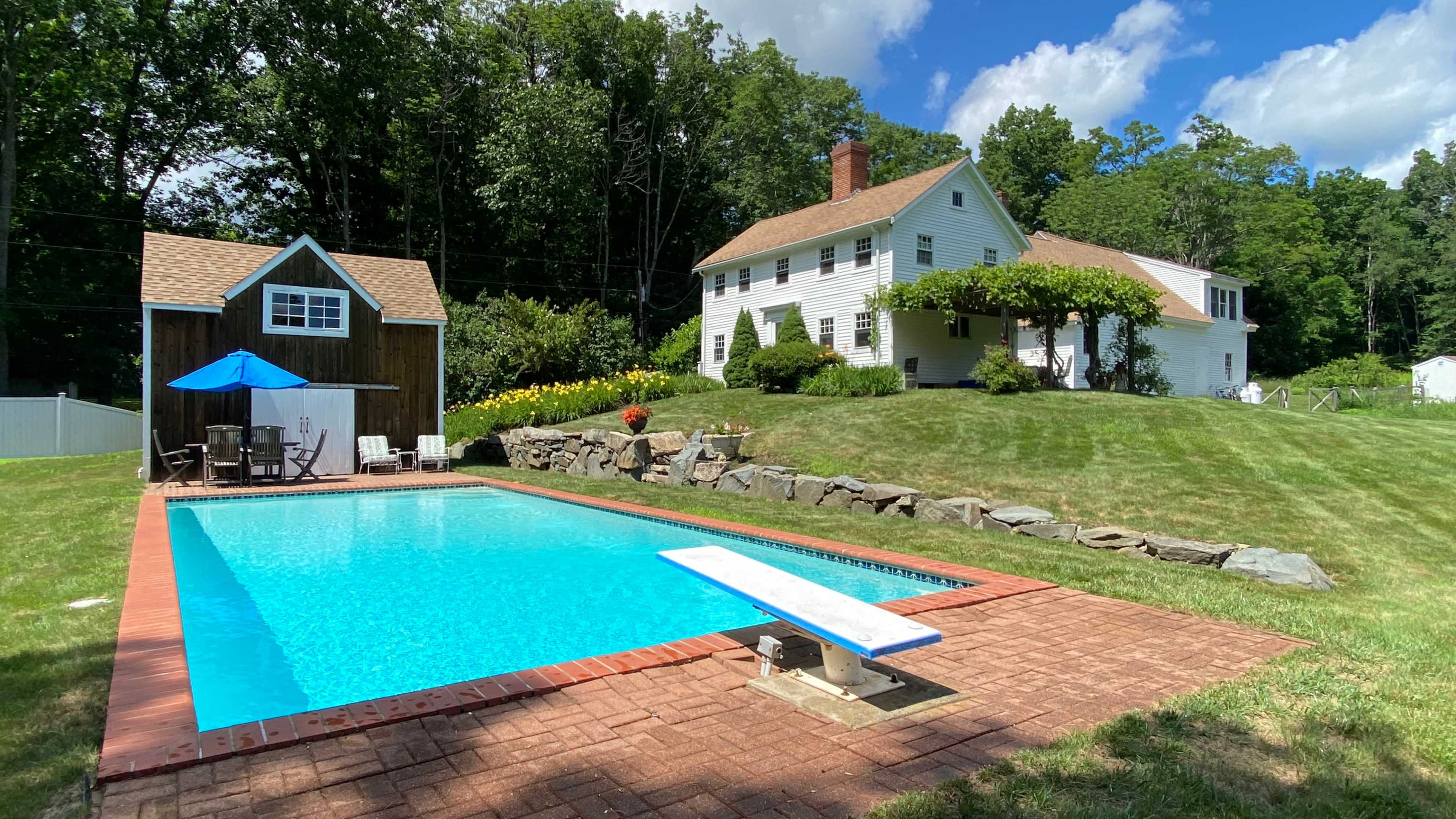 A swimming pool with a diving board is situated in a grassy backyard next to a white two-story house surrounded by trees.