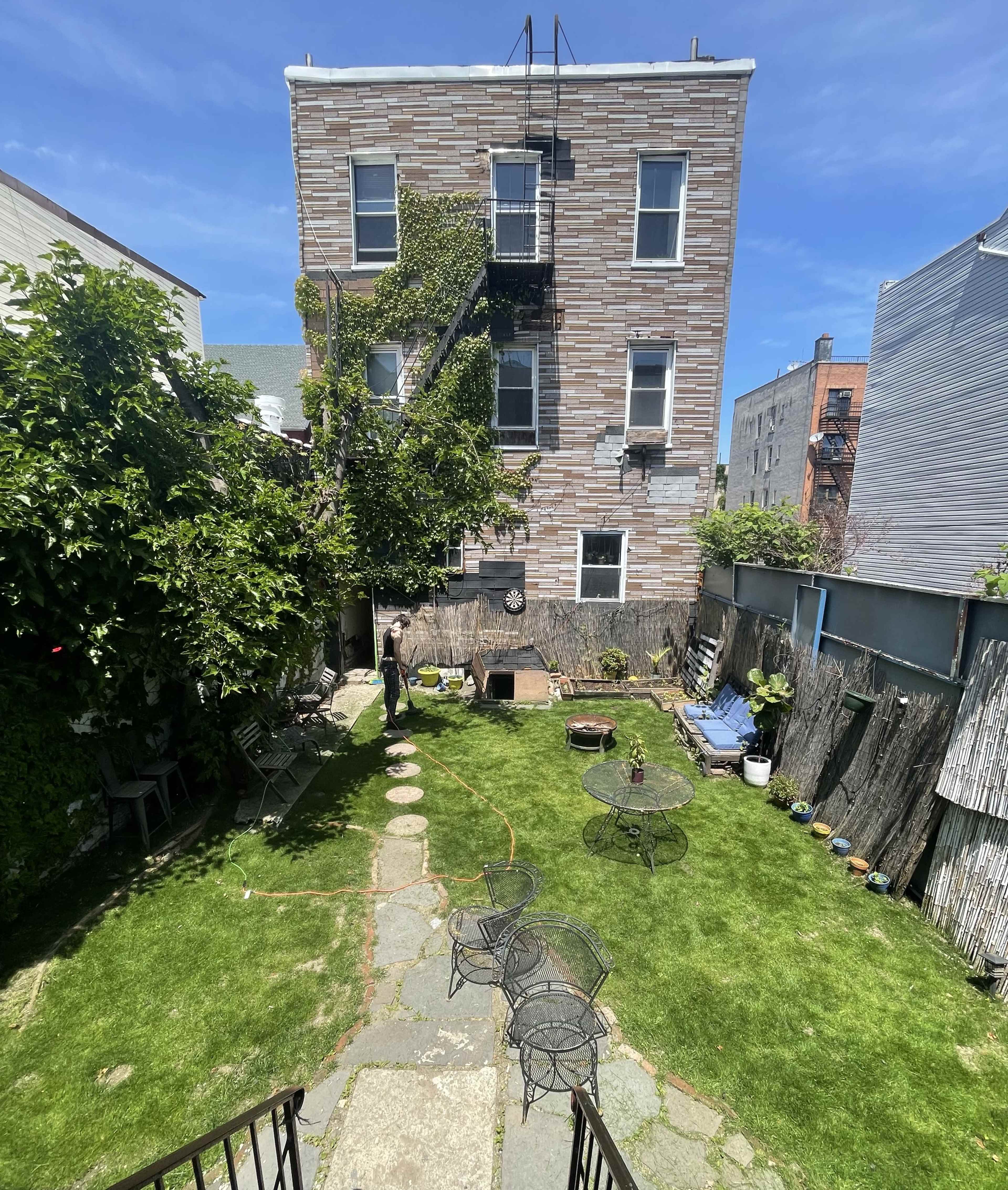 A garden area features a grassy lawn, stone path, and metal furniture, surrounded by a three-story brick building and various plants.